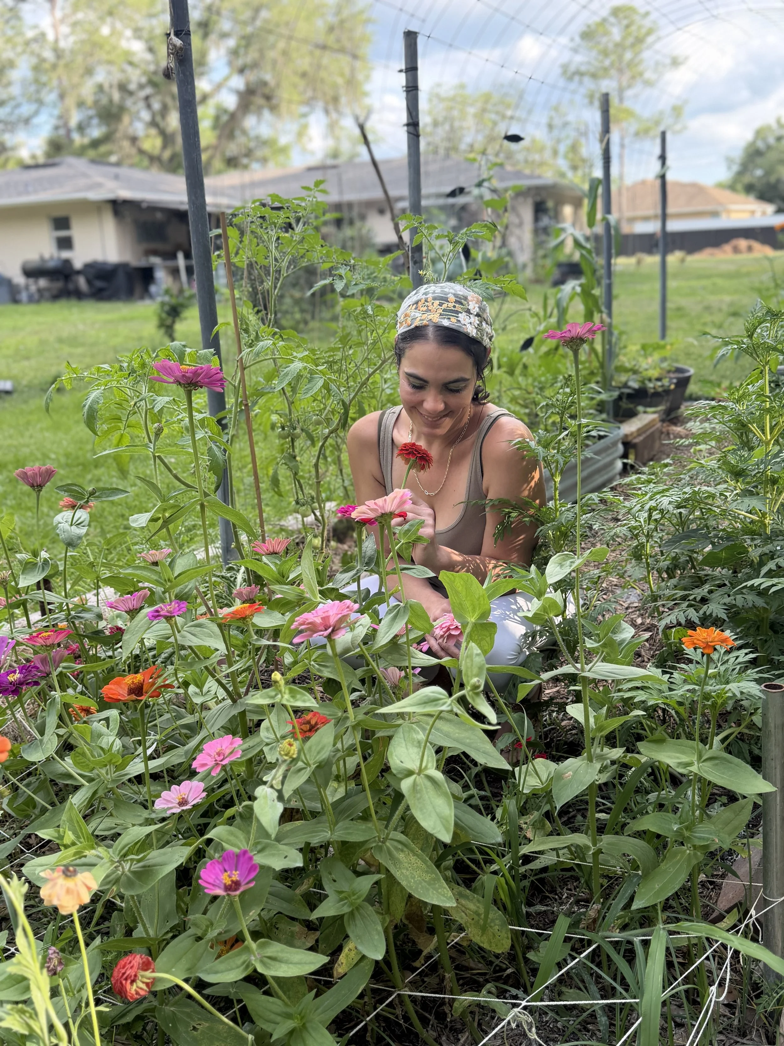 A woman is gardening in her backyard, surrounded by blooming pink, orange, and red flowers, with a house and trees in the background, during daytime. Owner of Bode Bars, in Lakeland,Fl. or online bodebars.com