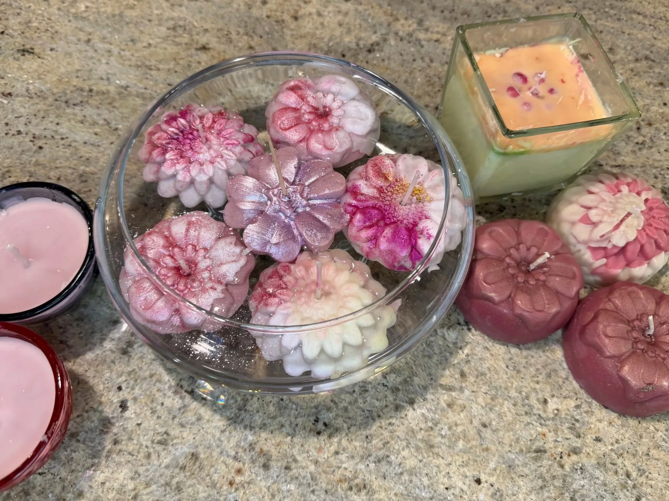 Ice candles shaped like flowers, in shades of pink and white, displayed in a glass bowl on a granite countertop, alongside a square glass container filled with colorful wax or candles.
