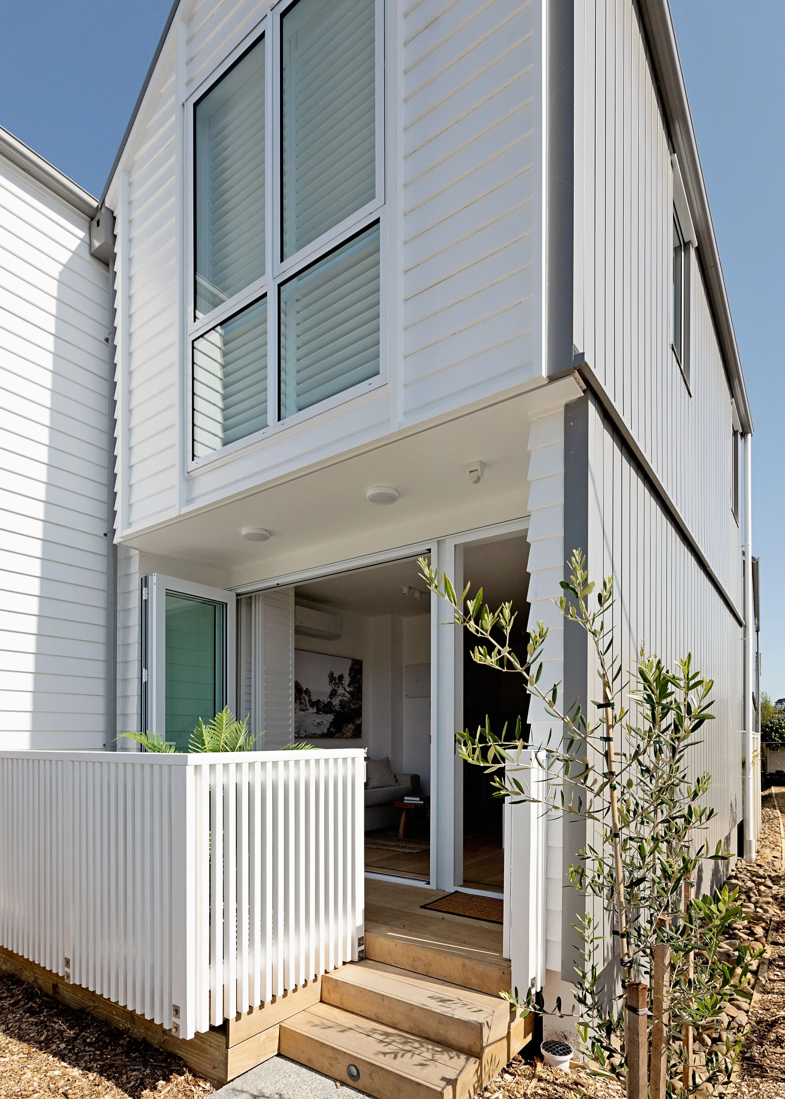 Close-up of a modern house exterior with white siding, large glass windows, a small porch with wooden steps, and a white picket railing, surrounded by some plants and rocks.