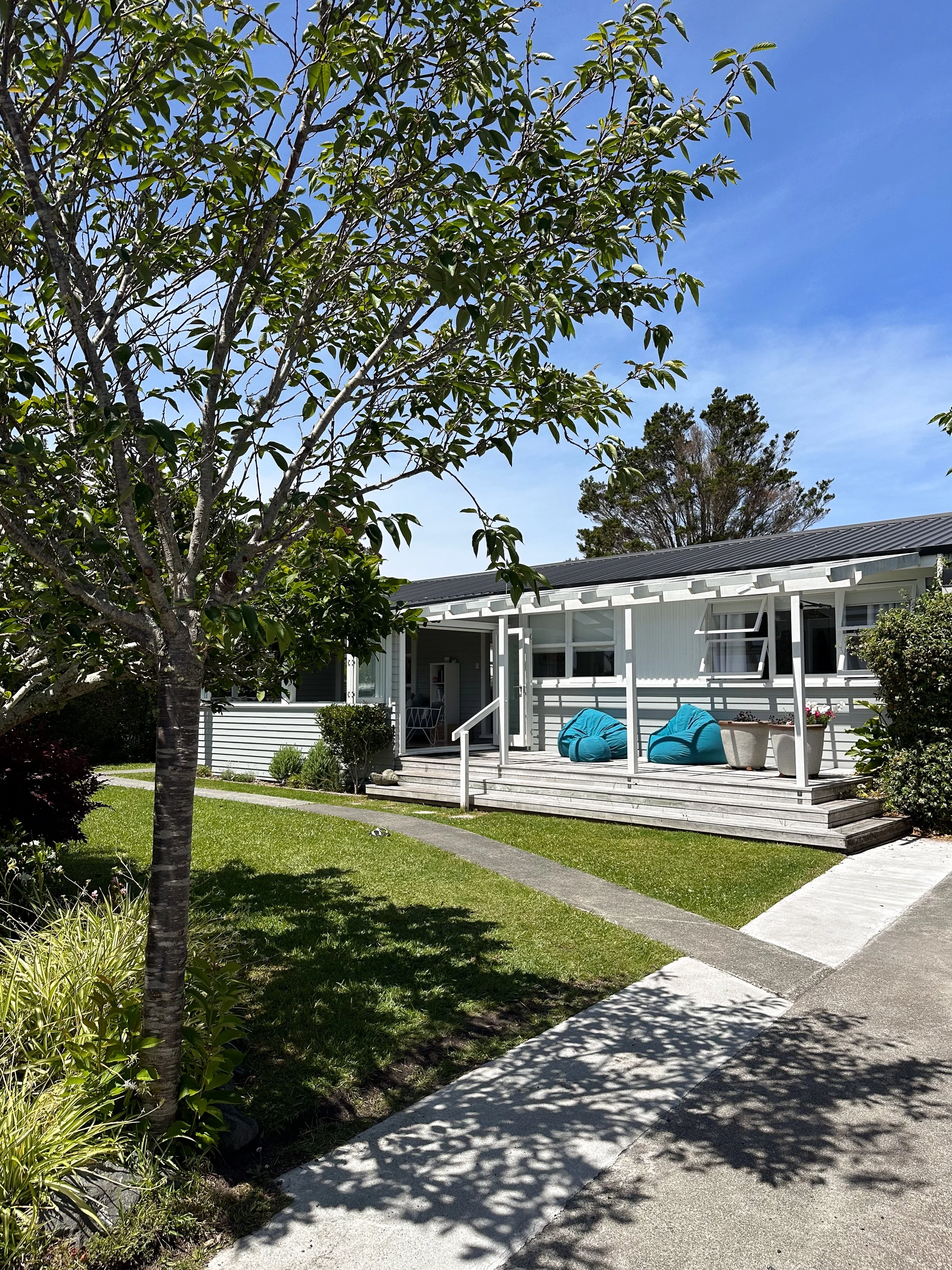A backyard scene with a tree on the left and a house with a porch on the right. The porch has blue bean bag chairs, a potted plant, and a small set of stairs leading to a grassy lawn with a concrete walking path. The sky is blue with some trees in the background.