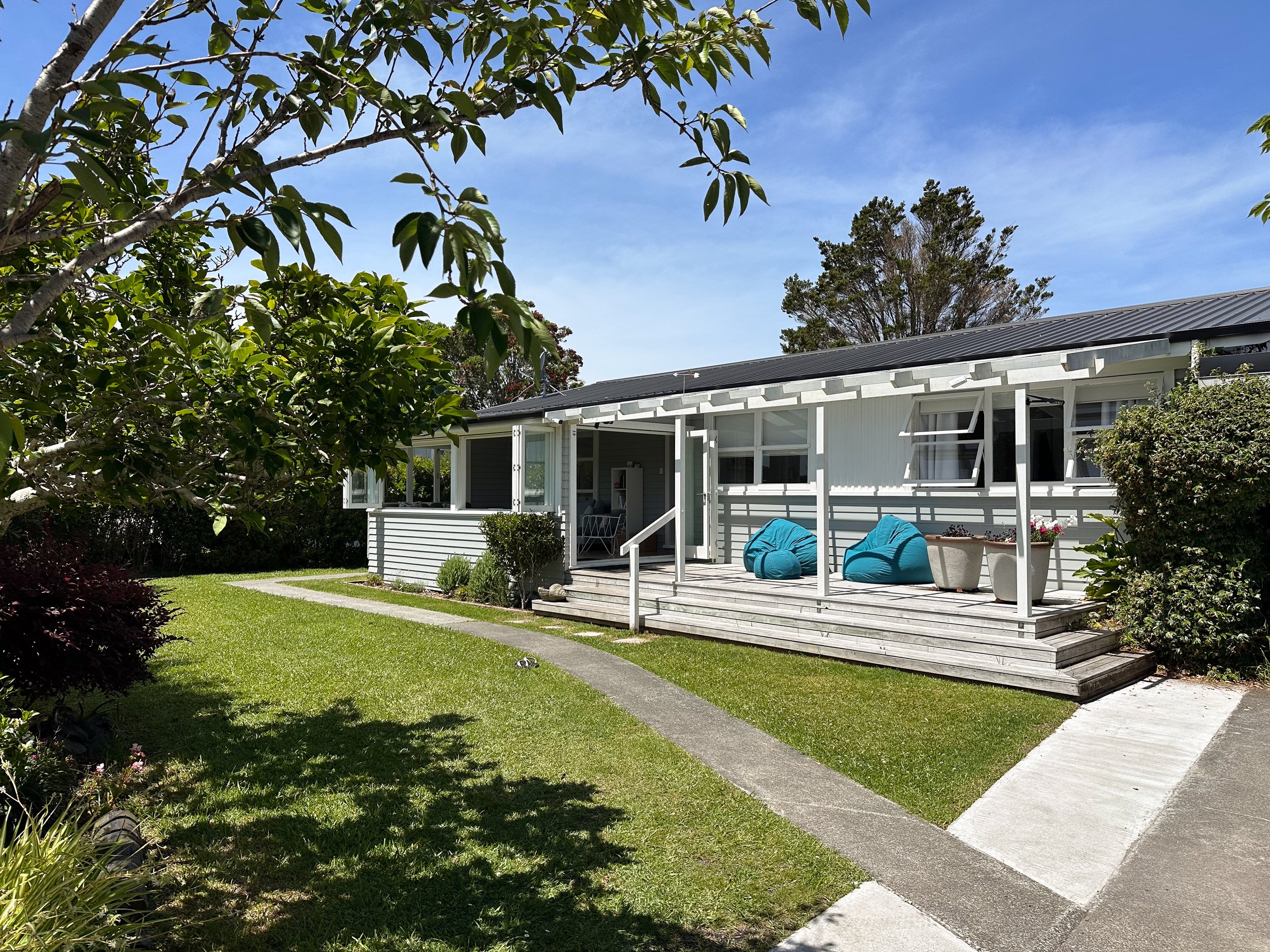 A backyard patio area with a white house, outdoor seating including blue chairs, a small table, potted plants, and lush green grass surrounded by trees and bushes under a clear blue sky.