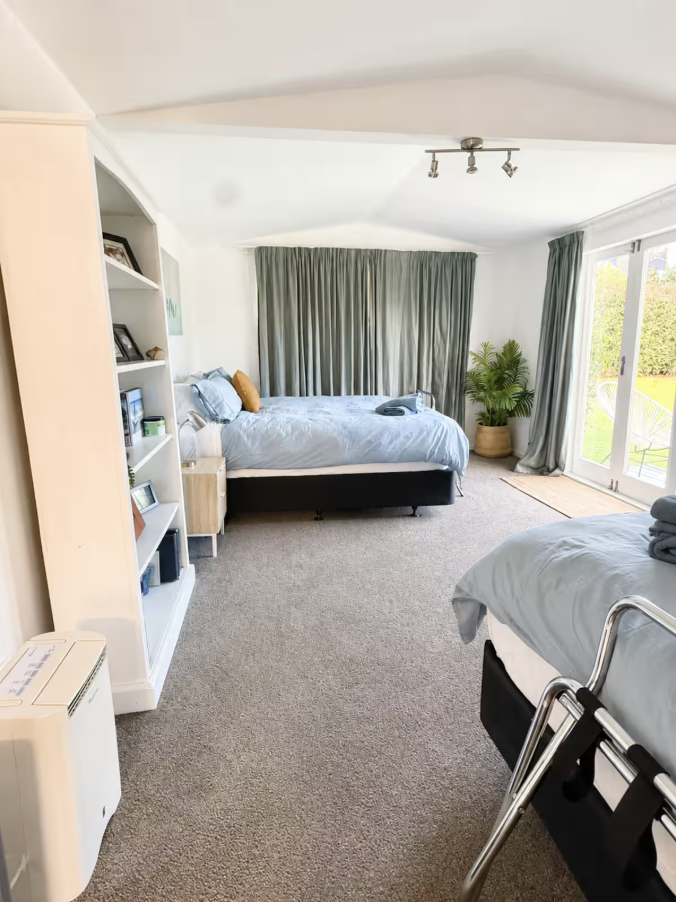 A bright bedroom with two beds, one near a sliding glass door, and a green plant in a pot. There is a bookshelf on the left side, and curtains behind the bed in the background. The floor is carpeted.