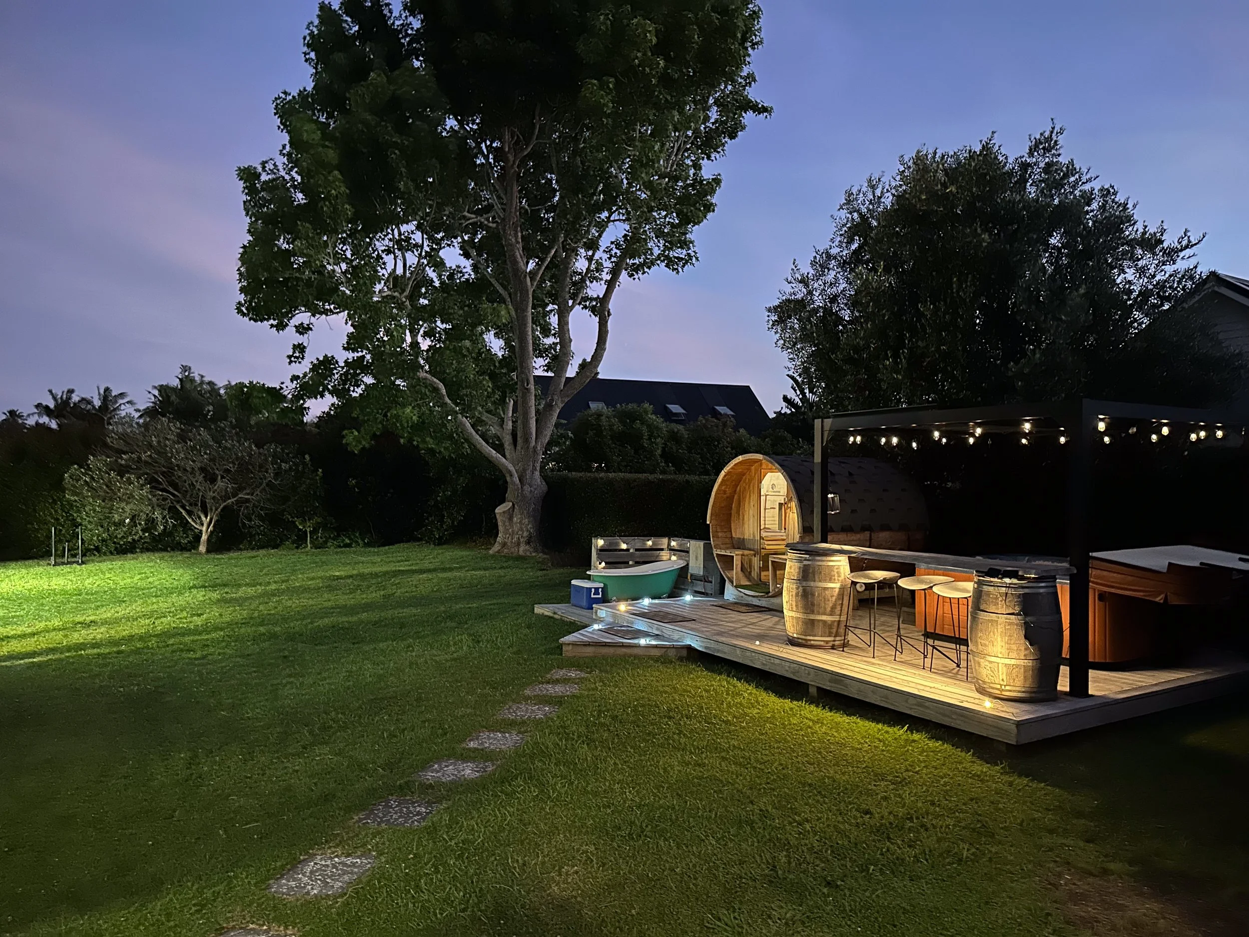 A backyard at dusk with a large tree, a wooden deck with string lights, barrels, and chairs, and a house in the background under a blue sky.