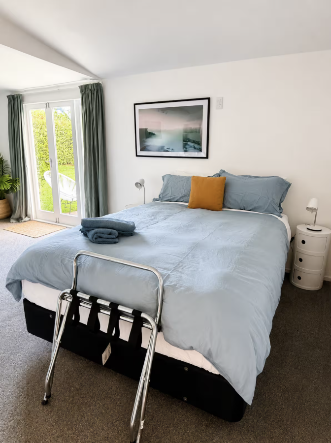 Bedroom with a bed covered in light blue sheets, a mustard-colored pillow, dark towels on the bed, and a framed landscape photograph on the white wall. There are sliding glass doors with green curtains leading outside, and a small white bedside table with a lamp.