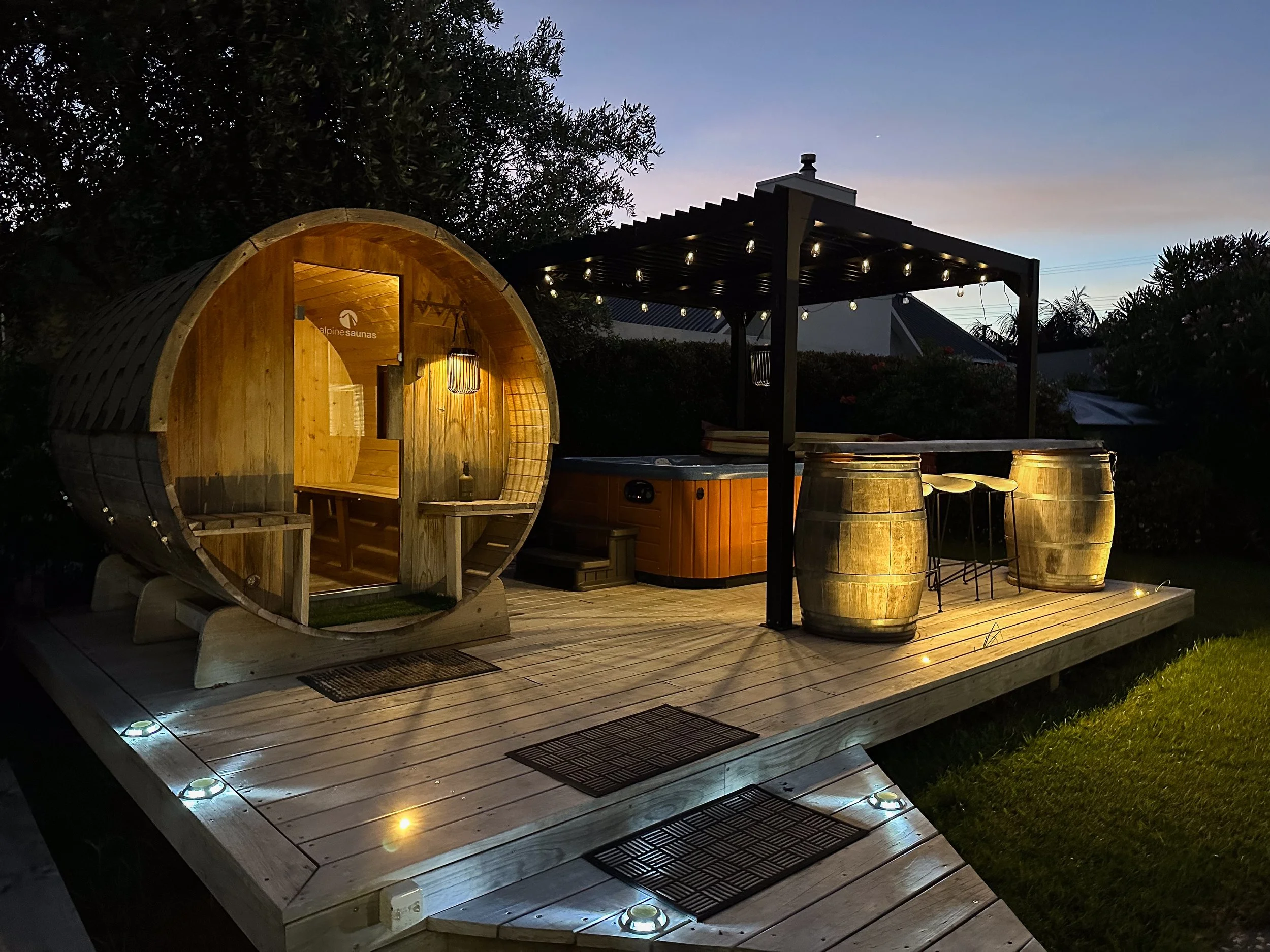 Outdoor wooden deck at dusk with a small wooden sauna shaped like a barrel, a hot tub, and string lights. The sauna has an open door with warm lighting inside. There are 2 barrels used as tables and stools, with string lights overhead and small ground lights illuminating the deck. Trees and a building are visible in the background.