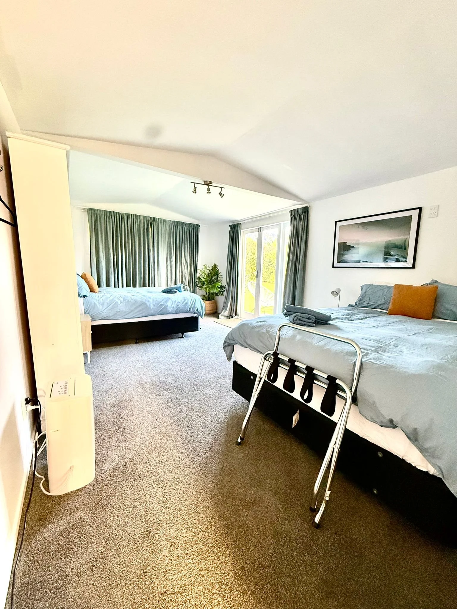 A room with two beds, one with a pillow and a phone, and the other with a gray blanket and a yellow pillow. There is a plant near the window, dark green curtains, and artwork on the wall. The room has carpeted flooring and a sliding glass door leading outside.