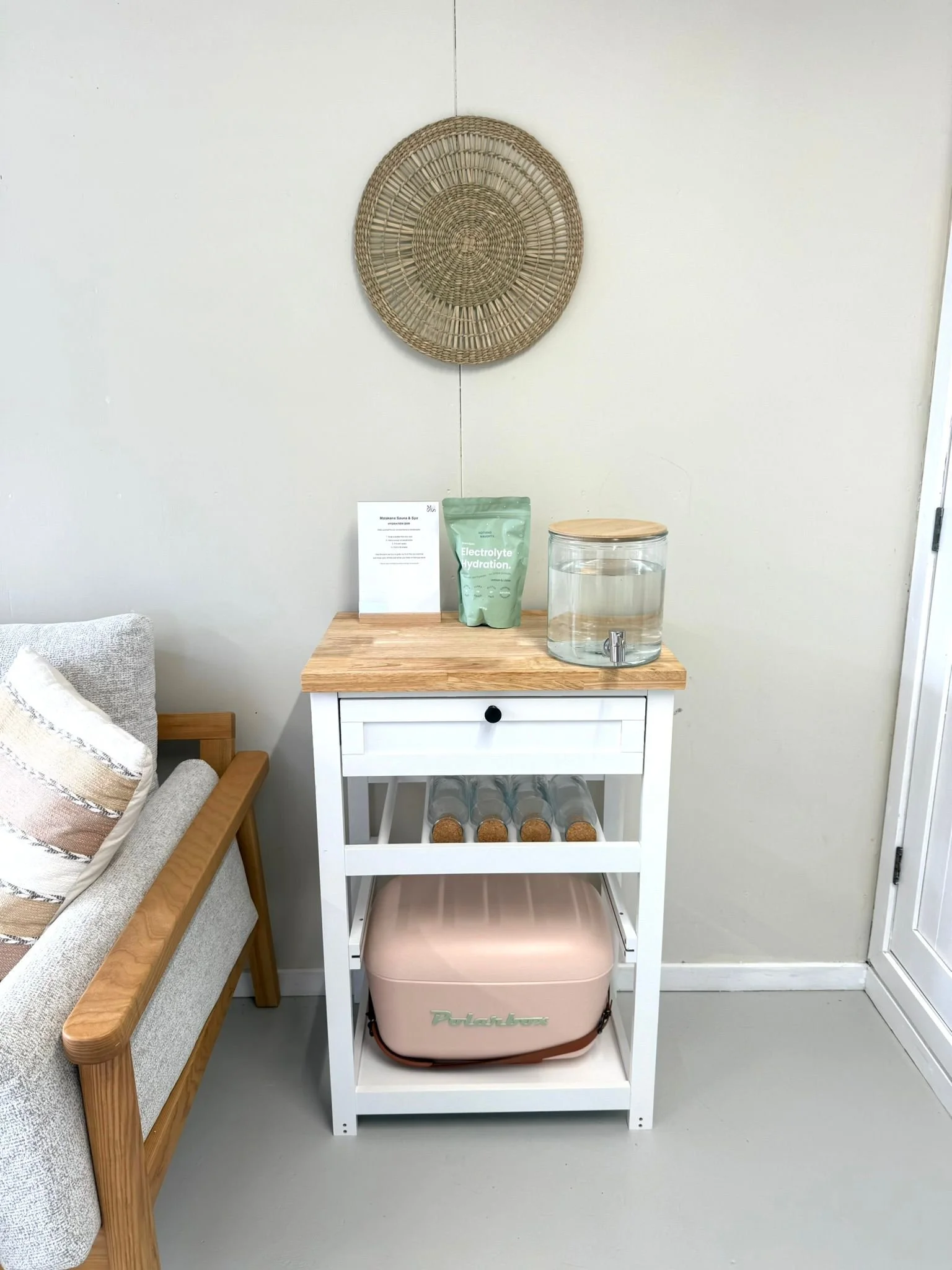A white three-tiered cart on wheels with a pink cooler bag underneath, a glass water dispenser with a wooden lid, and a green electrolyte hydration bag on top, positioned against a plain wall with a woven wall hanging above.