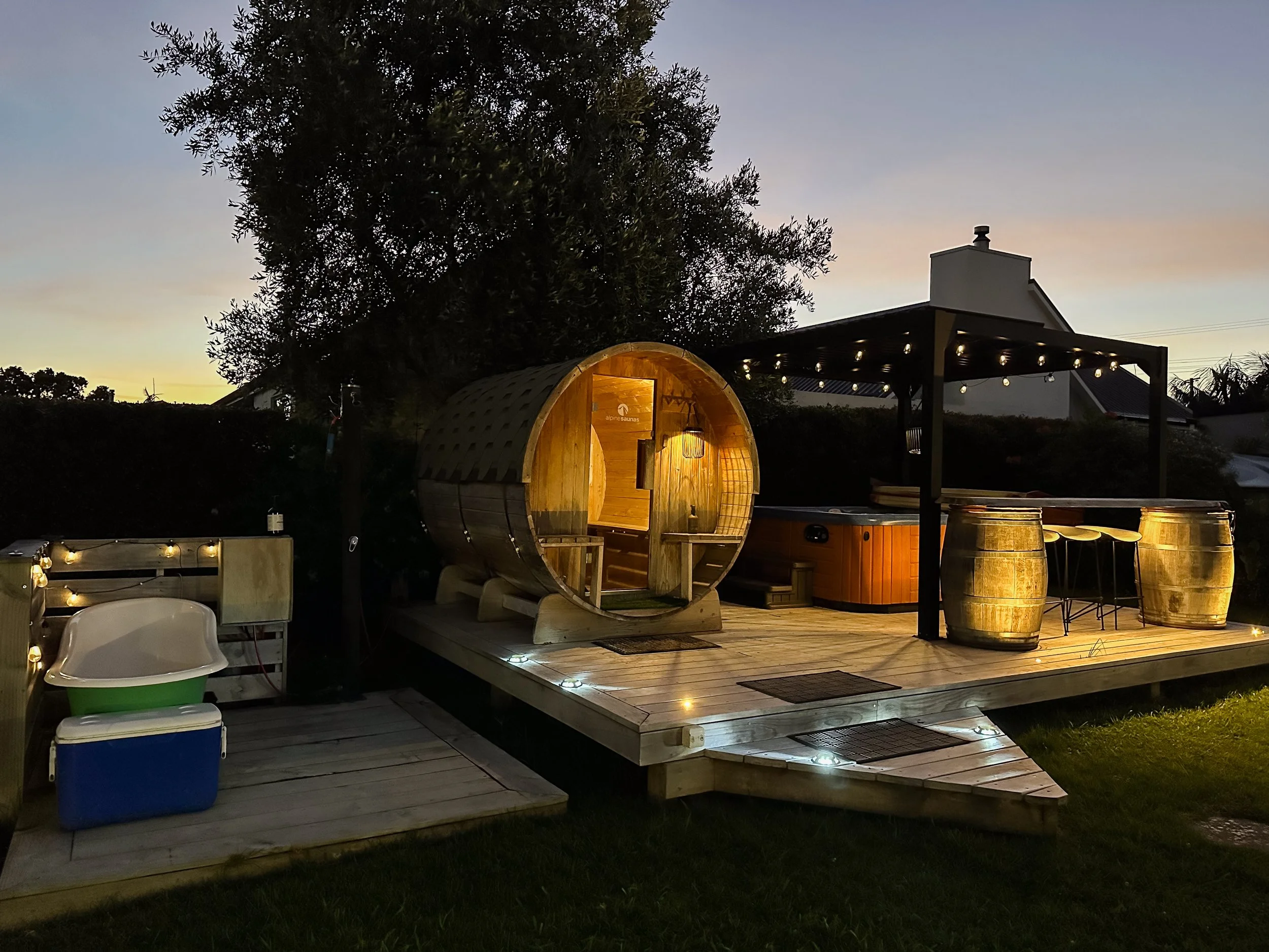 A backyard patio during early evening with a barrel sauna emitting warm light, string lights, a lounge chair, a small picnic table, and barrels used as tables, with a large tree and a house with a chimney in the background.