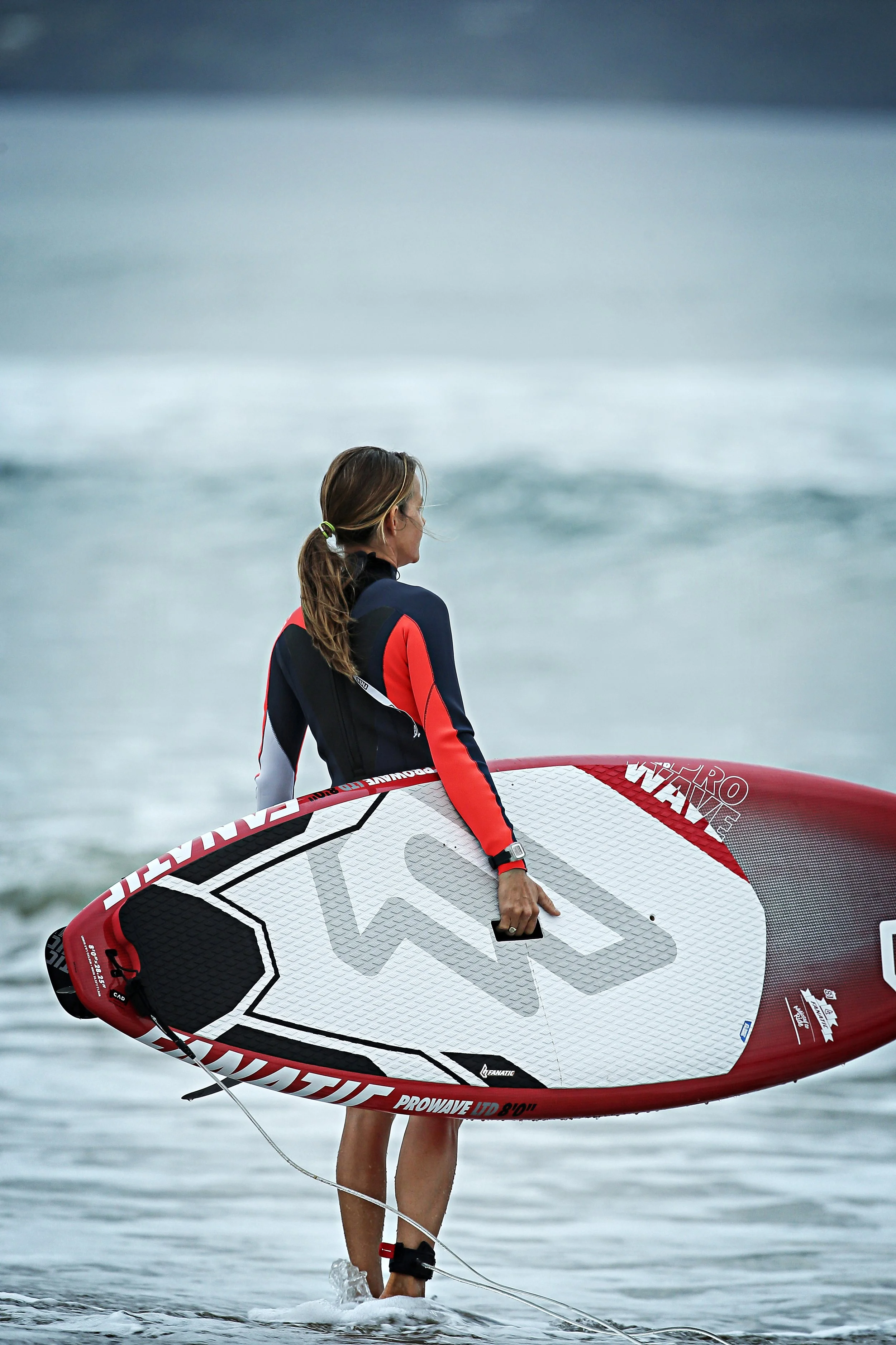 A woman standing in seawater holding a red and white stand-up paddleboard. She is wearing a black and red wetsuit, with her hair in a ponytail, and is overlooking the ocean.