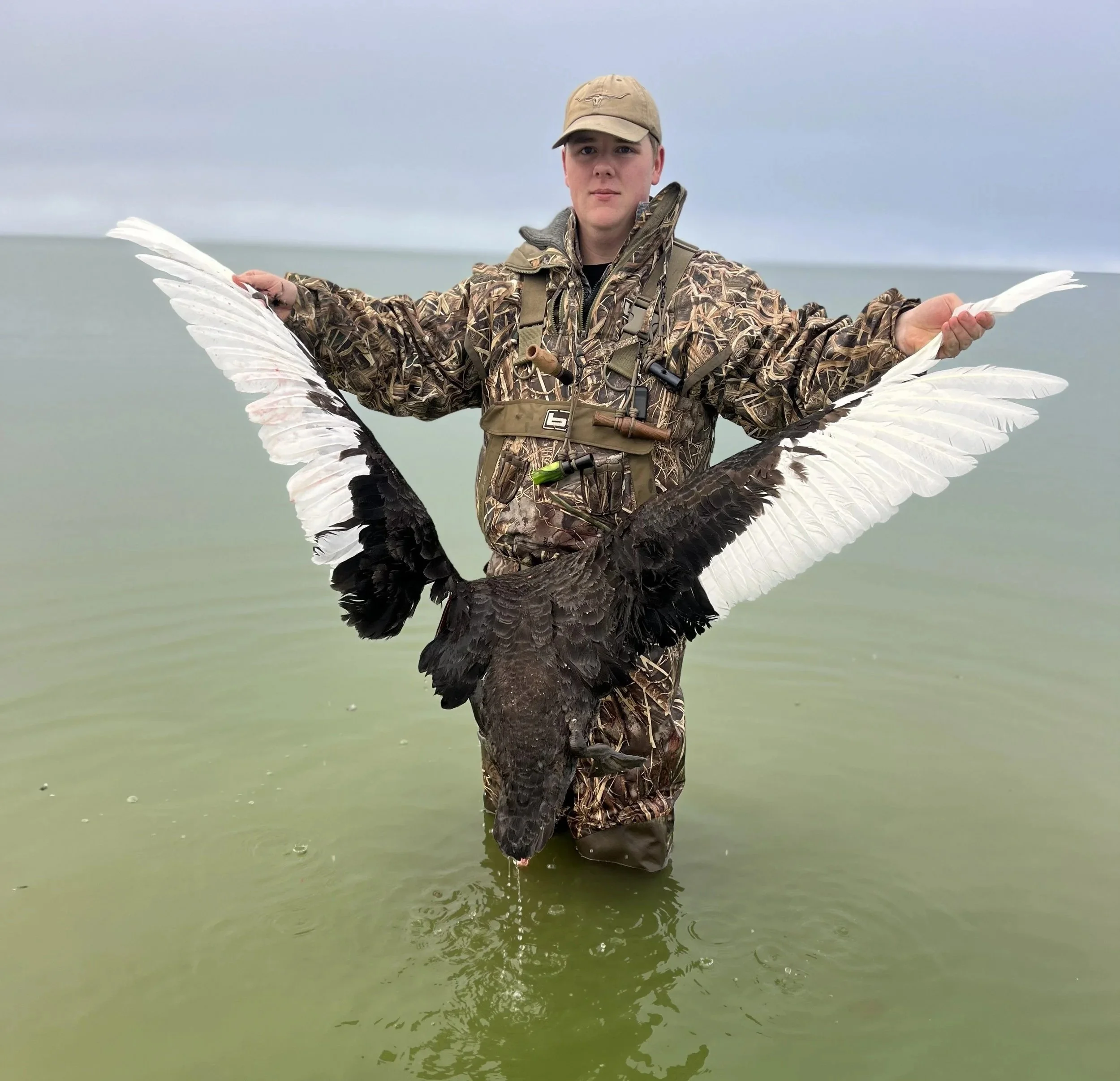 A person in camouflage clothing and a tan cap is standing in water, holding a large bird with black and white wings.