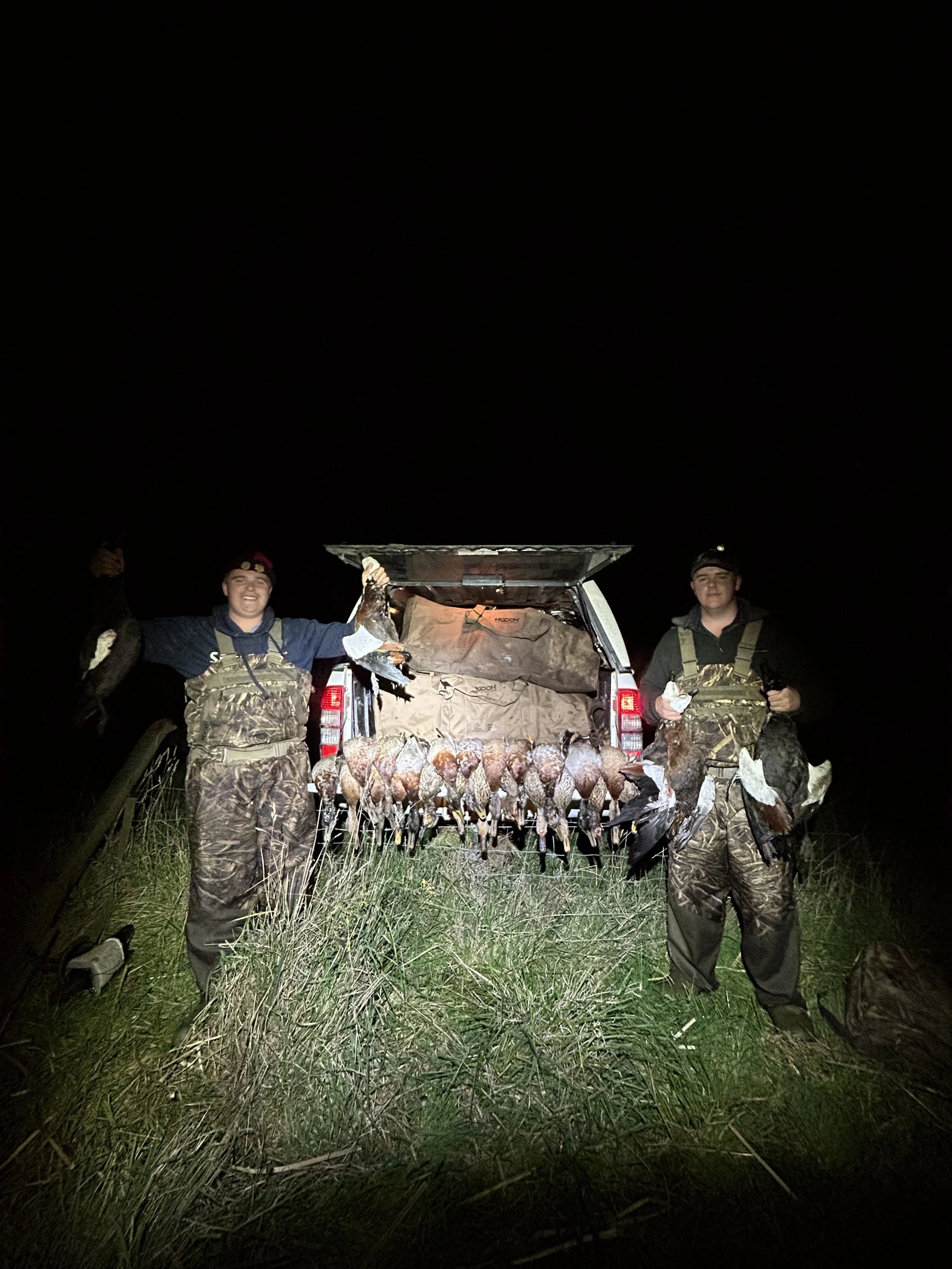 Two hunters in camouflage clothing display their hunting success, holding ducks in front of a truck with a load of ducks hanging from the open tailgate at night.