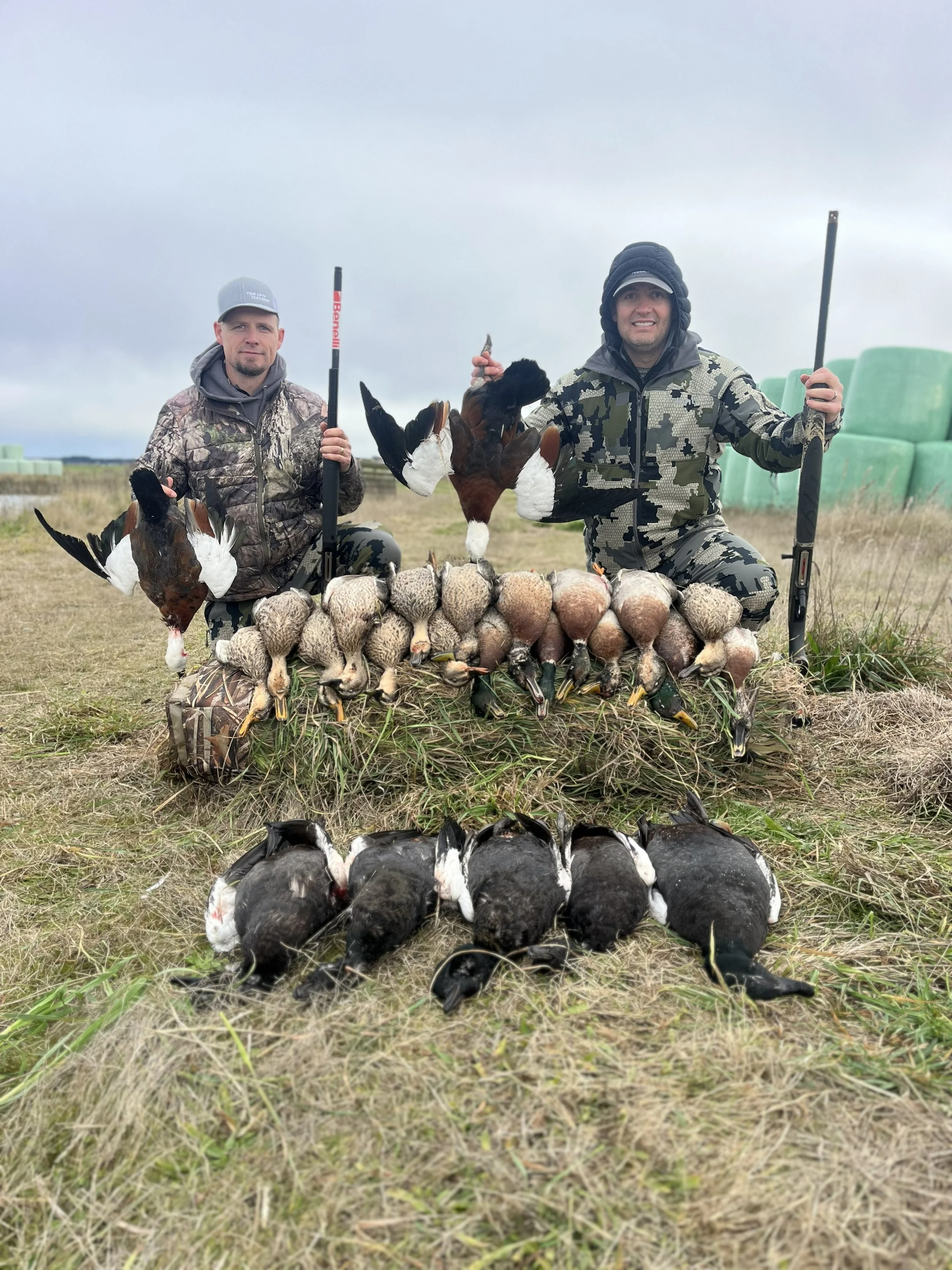 Two hunters kneeling on a field with their hunting guns, surrounded by a large collection of hunted game including ducks, pheasants, and other waterfowl, with hay bales in the background.
