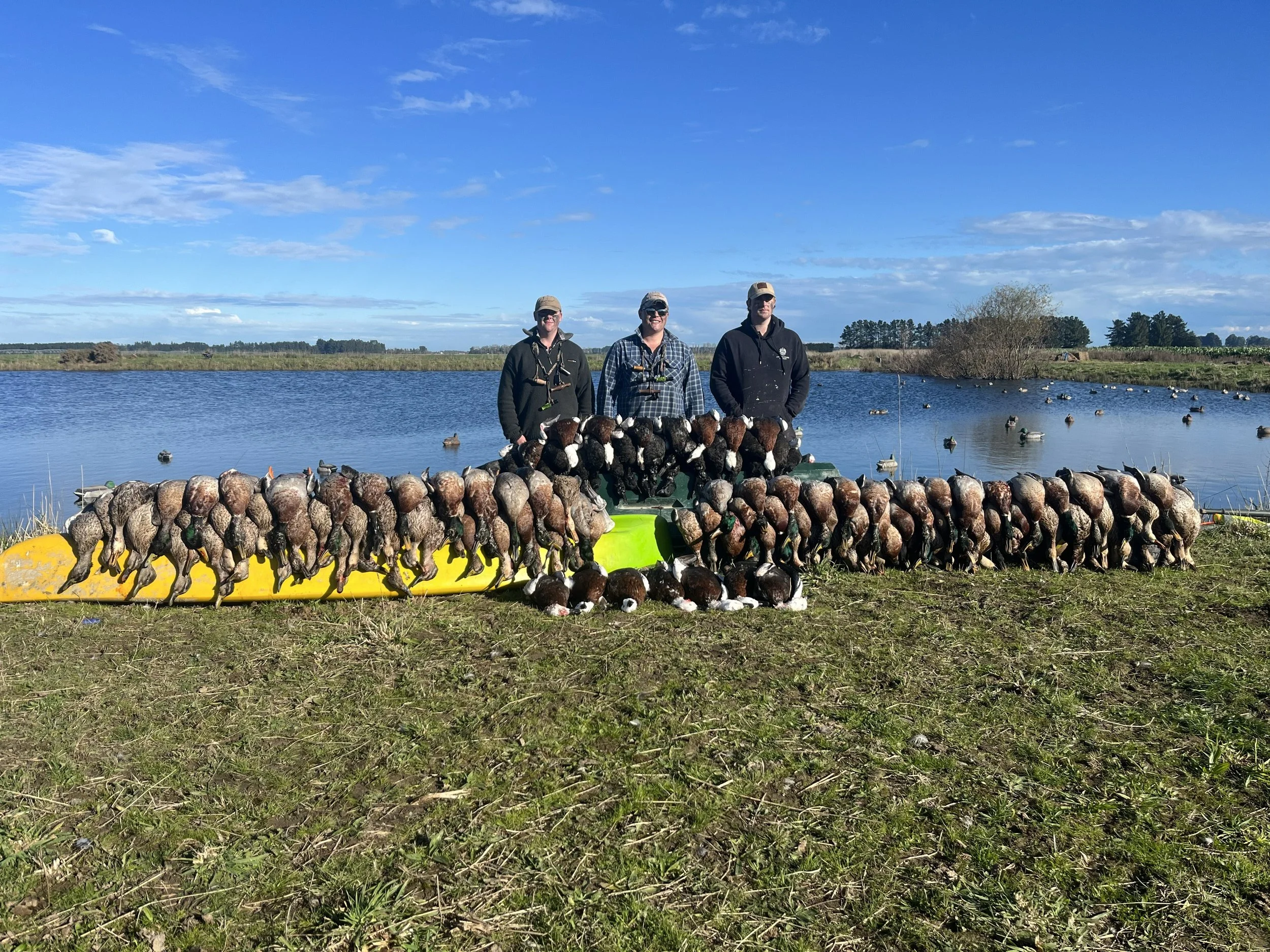 Three people stand on the shore of a lake behind a large array of hunted ducks laid out on a yellow and green watercraft, with the lake and a flock of ducks swimming in the background under a partly cloudy sky.
