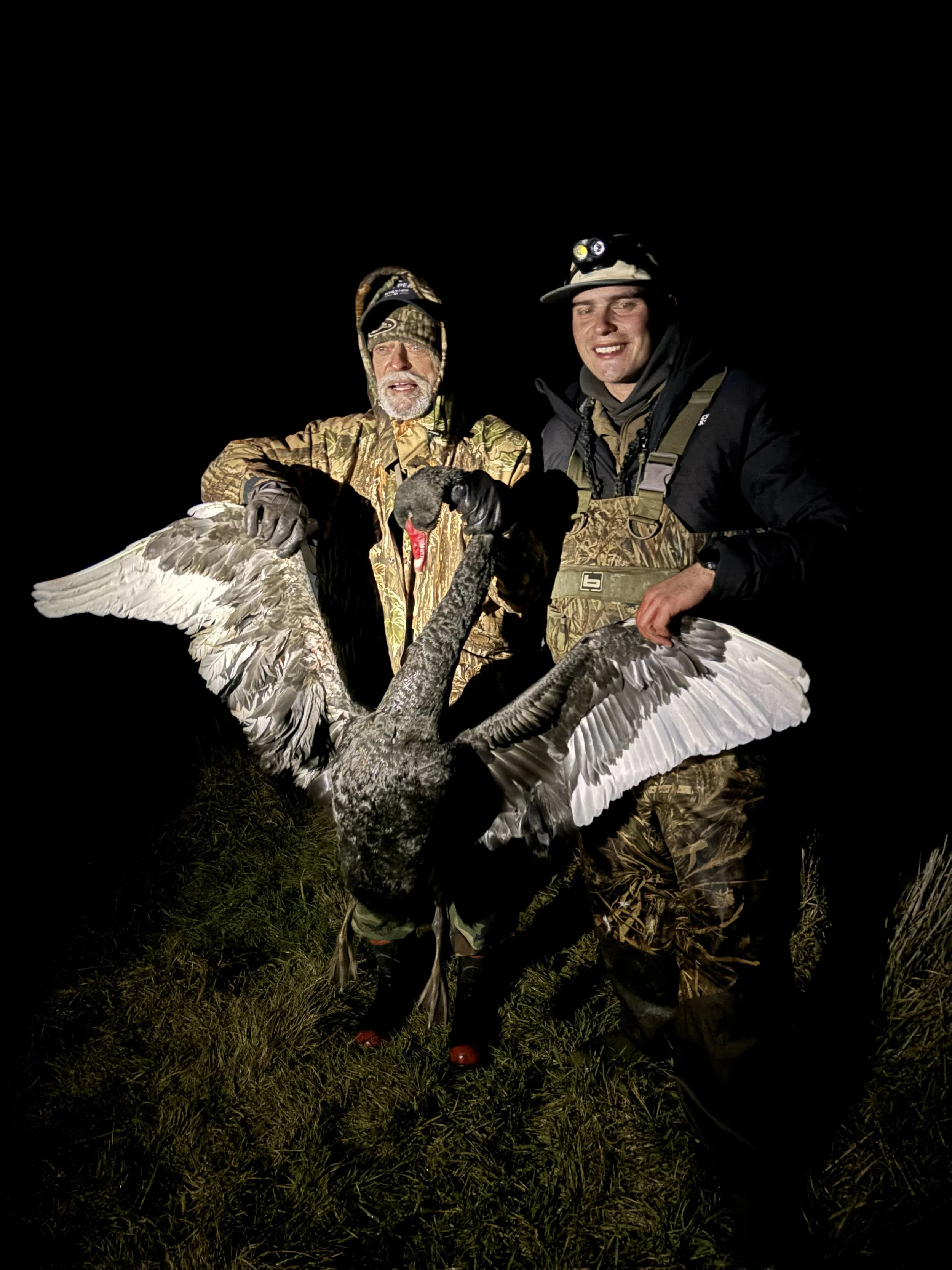 Two hunters in camouflage clothing at night holding a large dead crane with wings spread.