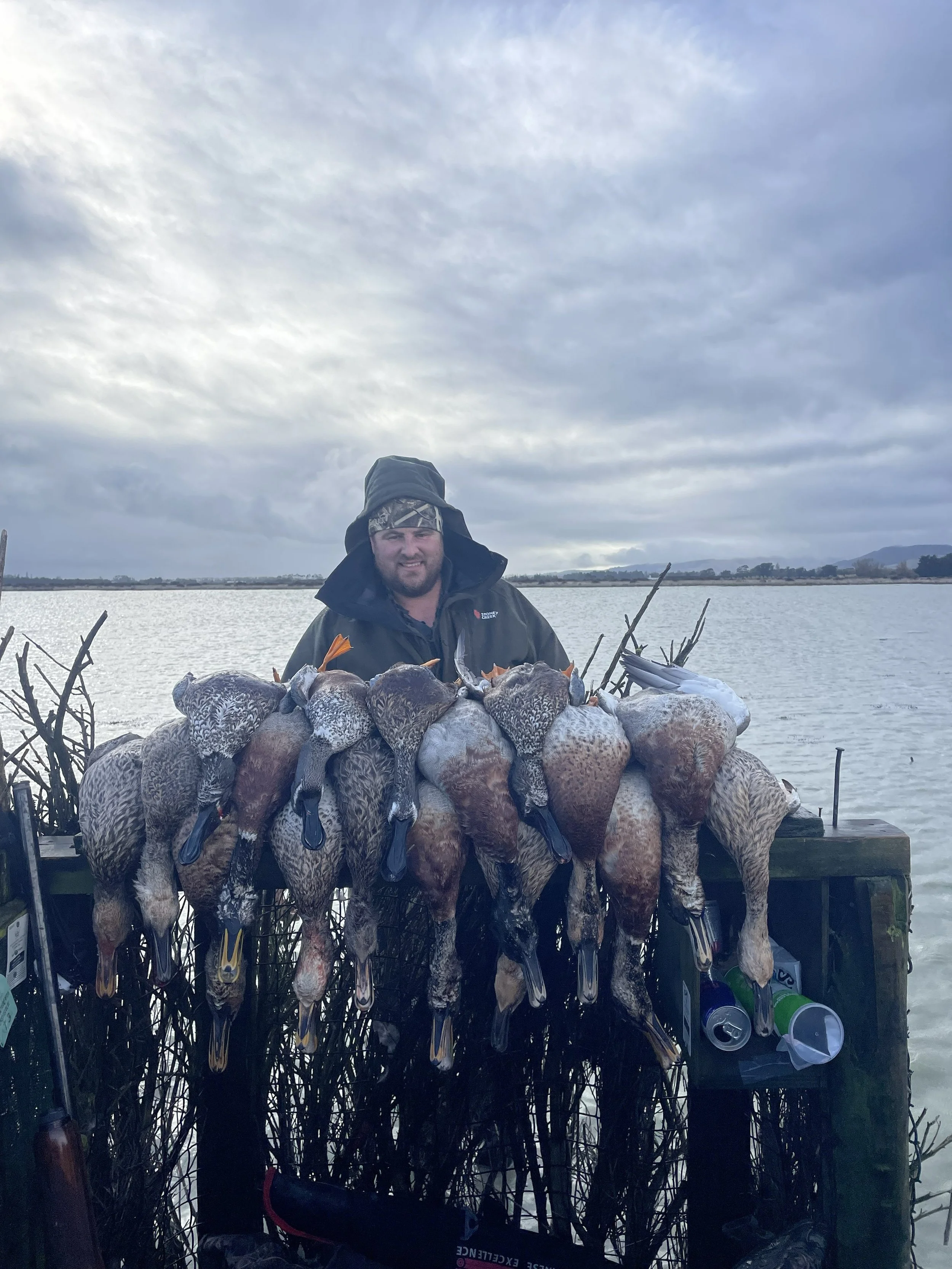 A man dressed in outdoor clothing standing behind a large number of ducks he has hunted, with a body of water and cloudy sky in the background.