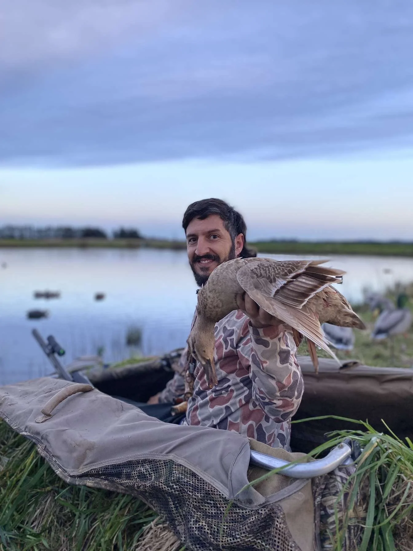 Man with a beard sitting by a lake holding a duck, with other ducks in the background, during sunset or dusk.