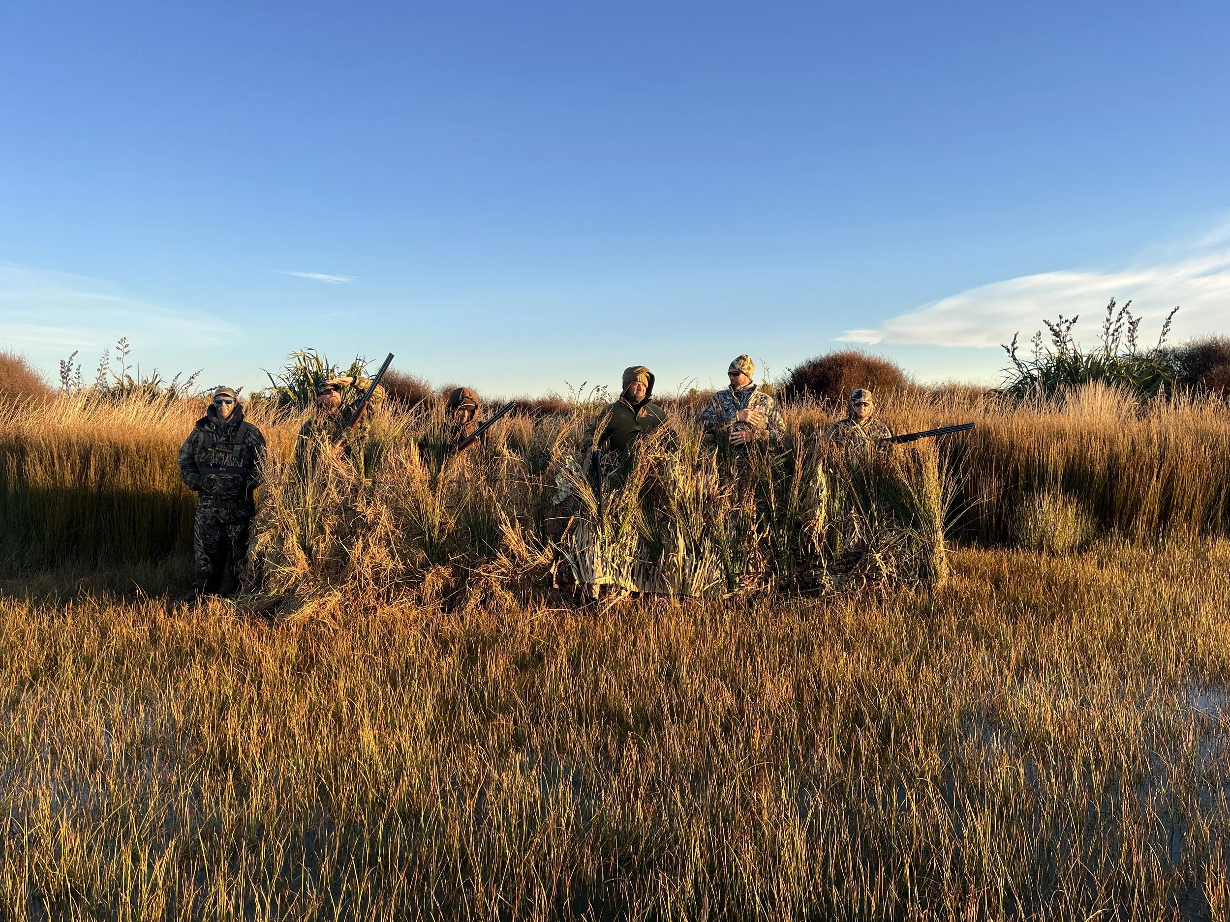 Six hunters standing among tall grass in a marsh during sunset, wearing camouflage and holding rifles.