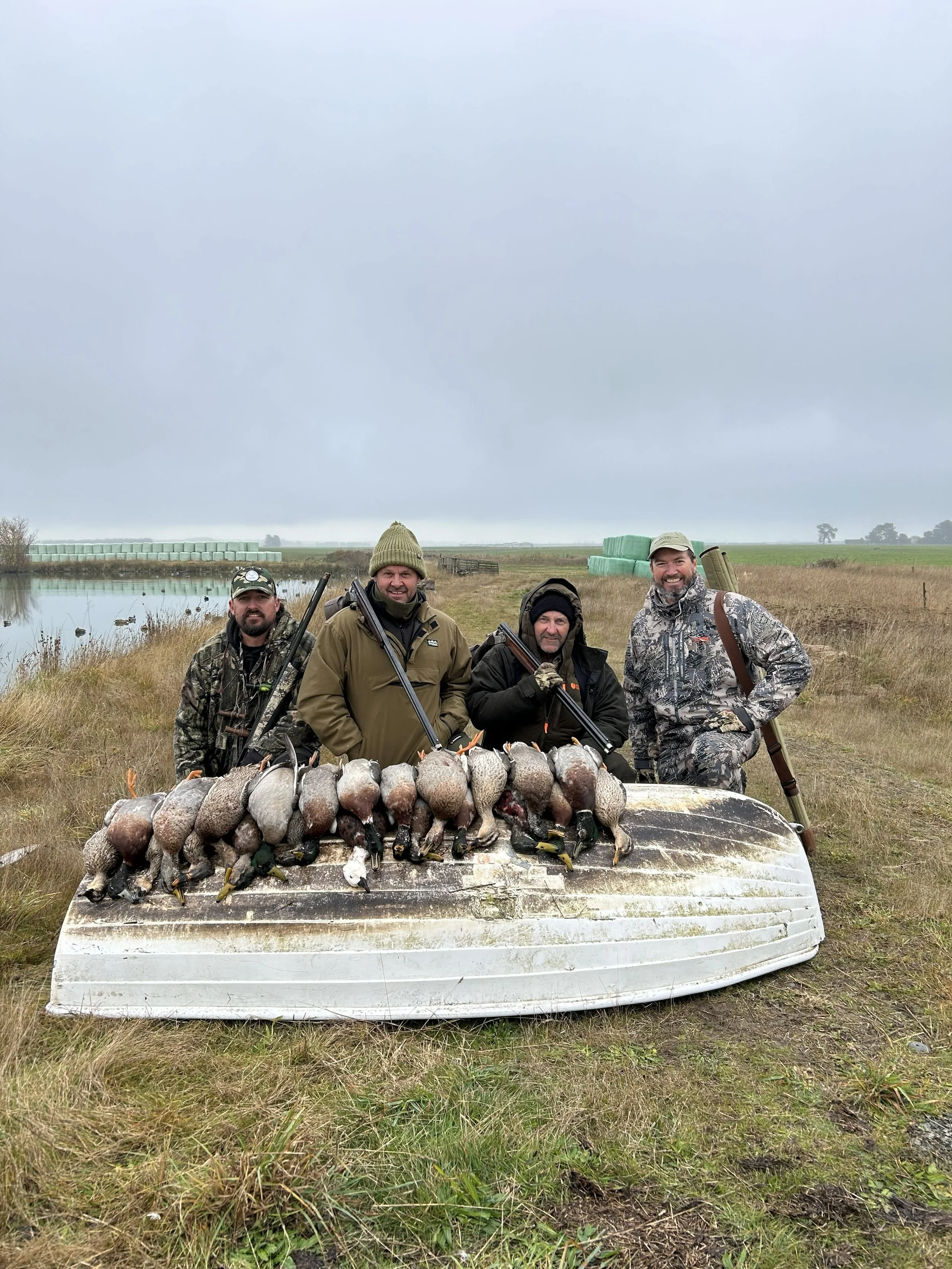 Four men standing behind a boat filled with ducks they hunted, in an open field near a body of water, on a cloudy day.