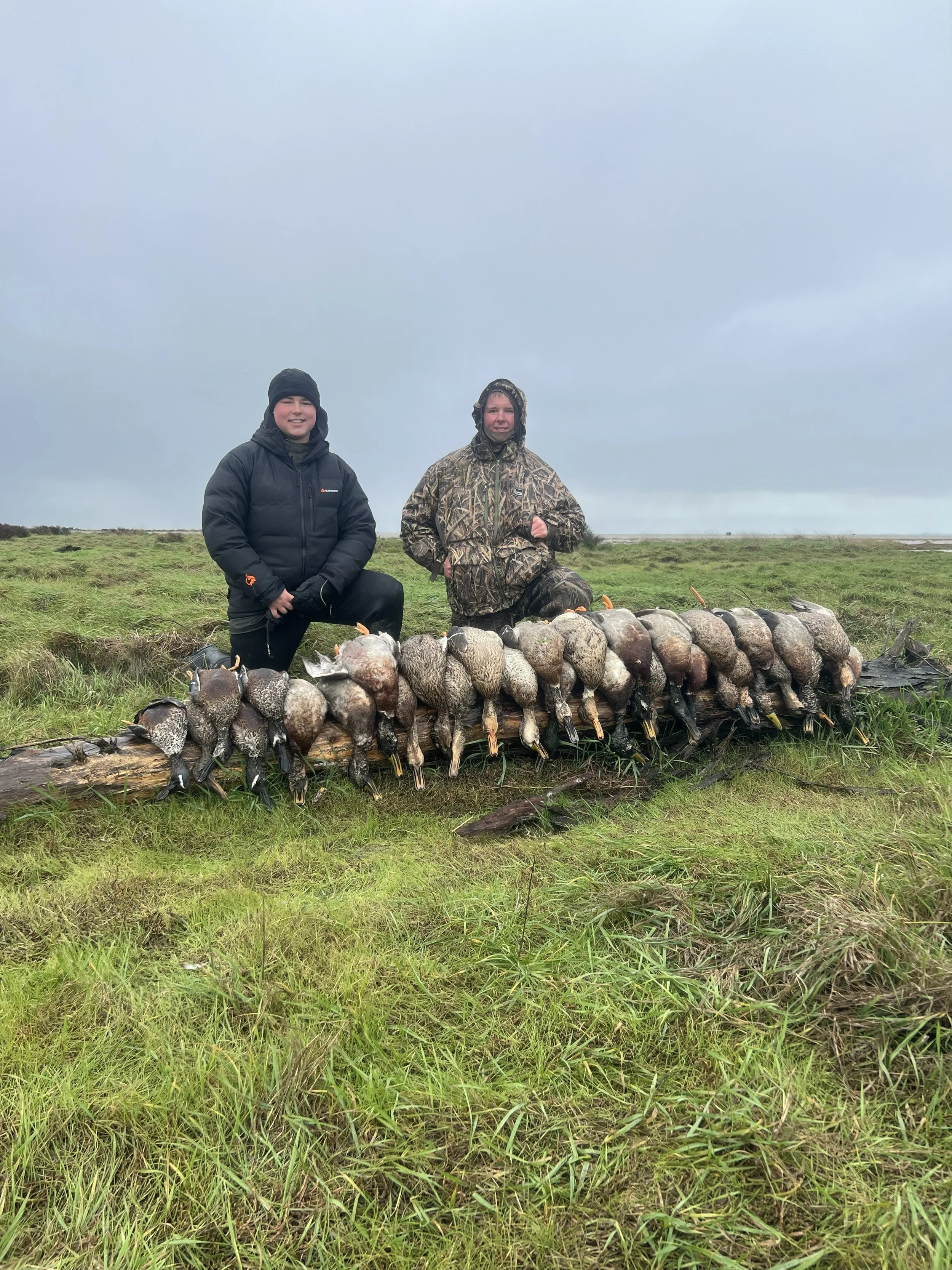 Two people dressed in outdoor hunting gear kneeling on each side of a row of dead ducks laid on a log, with a grassy field and cloudy sky in the background.