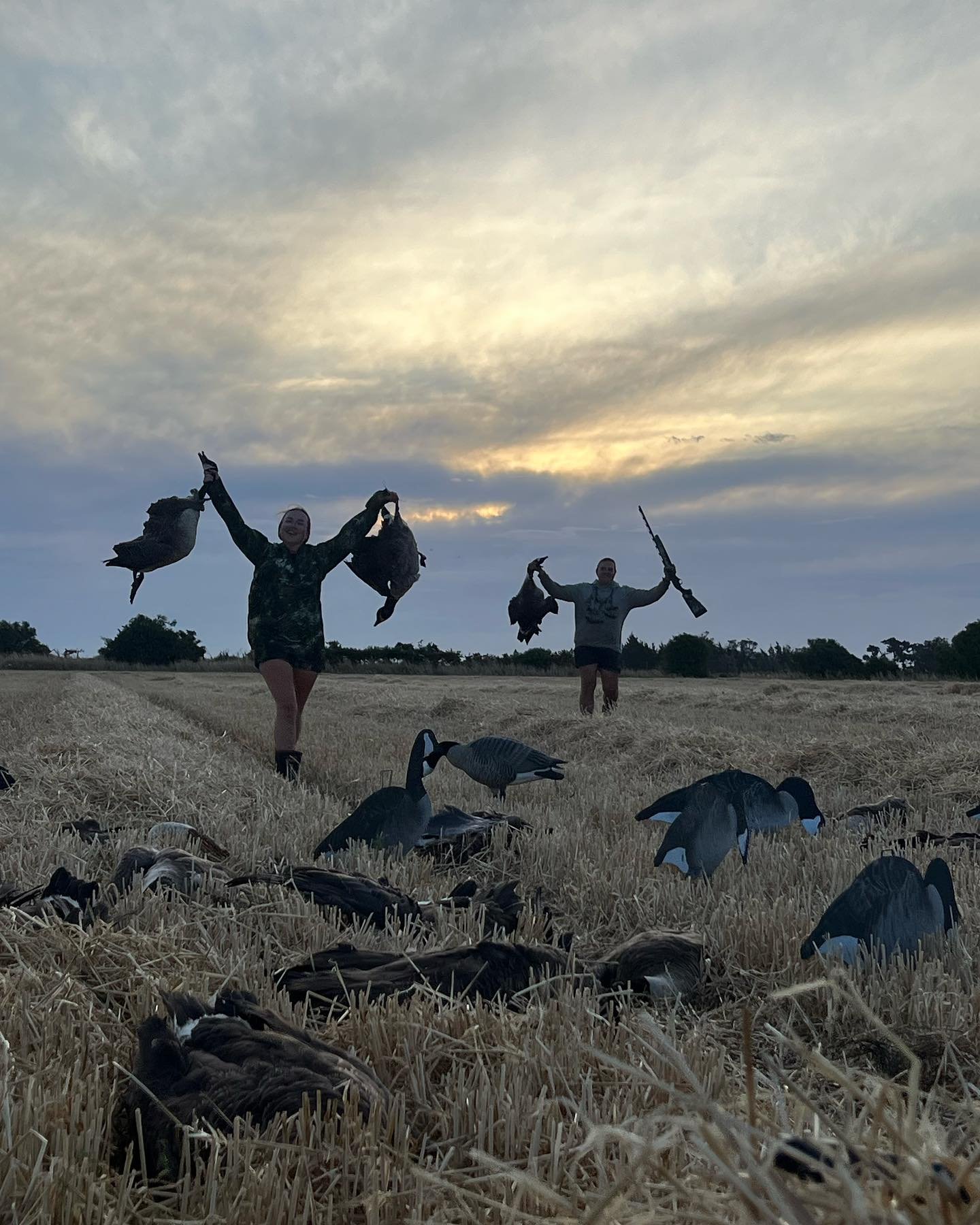 Two people in a field at sunset holding dead ducks, with additional ducks on the ground around them.