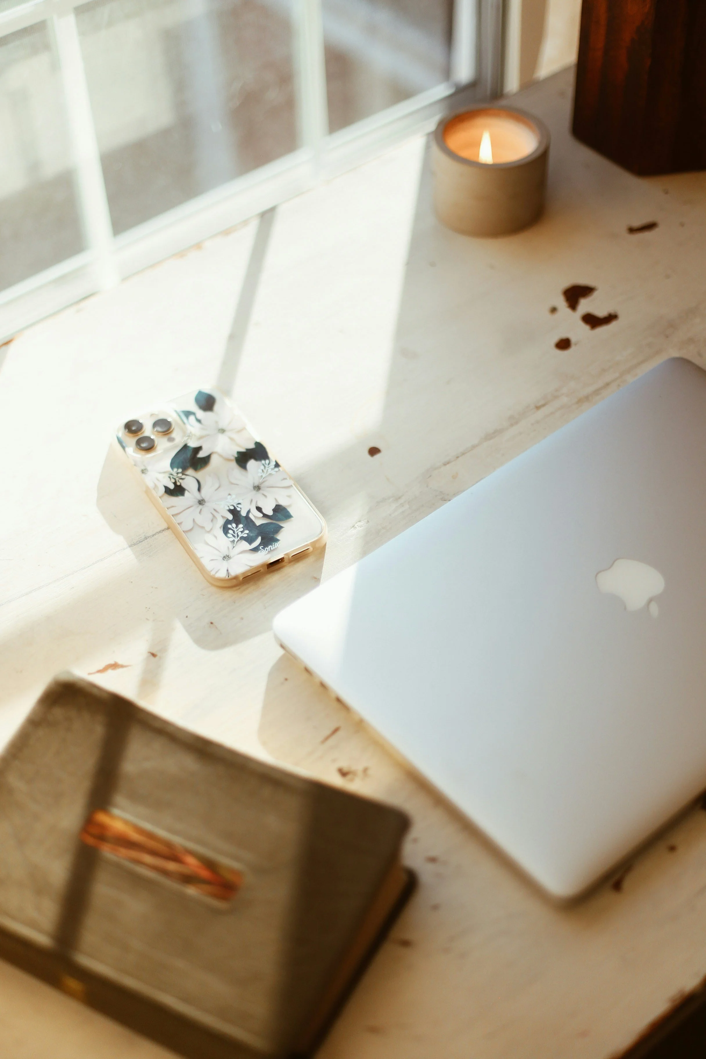 A small floral-patterned electronic device, a candle in a wooden holder, a silver laptop, and a metal jewelry box on a wooden table near a window with sunlight.
