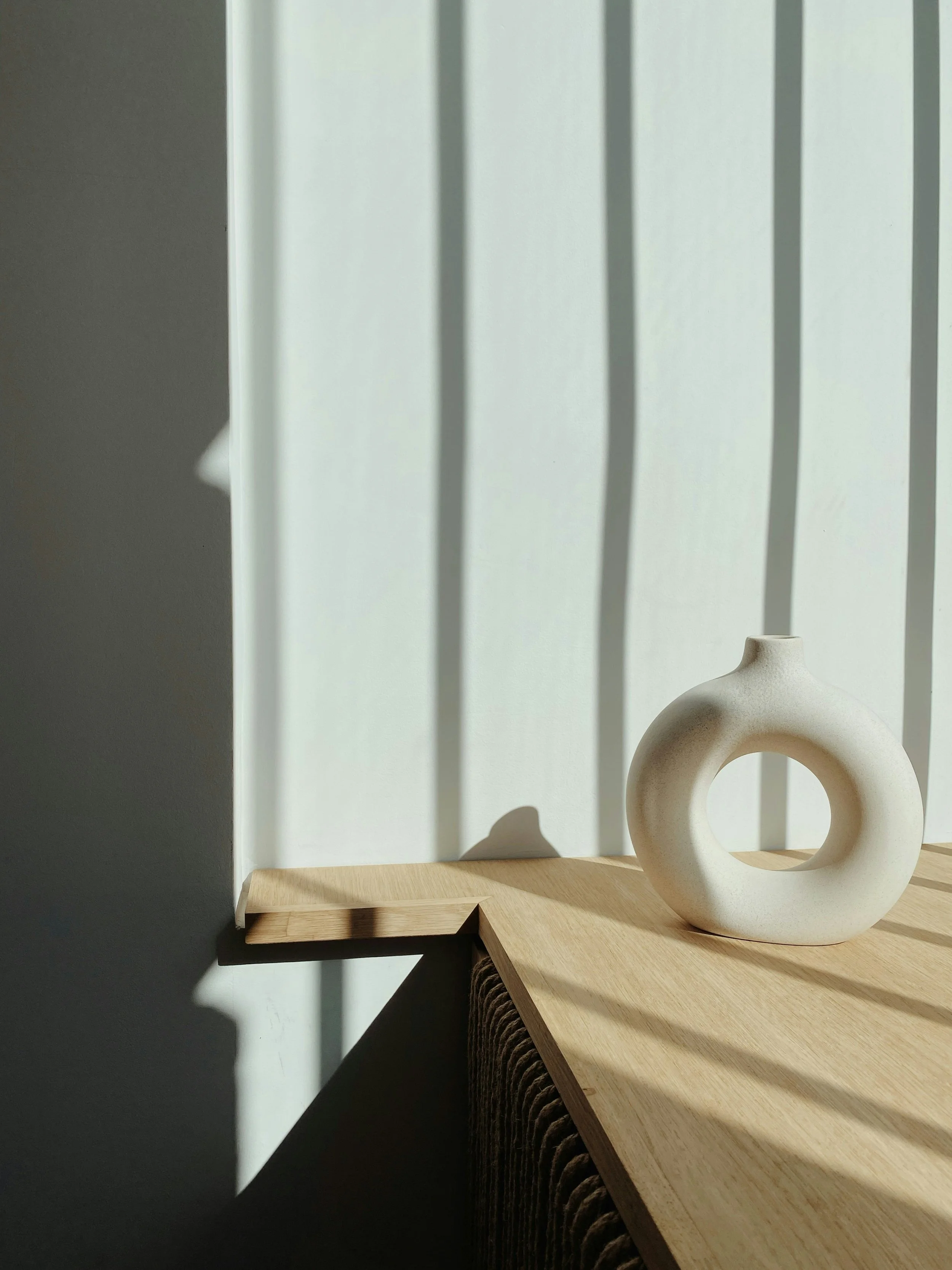 A minimalist white ceramic sculpture with a circular hole in the center, placed on a wooden surface next to a wall with vertical shadows.