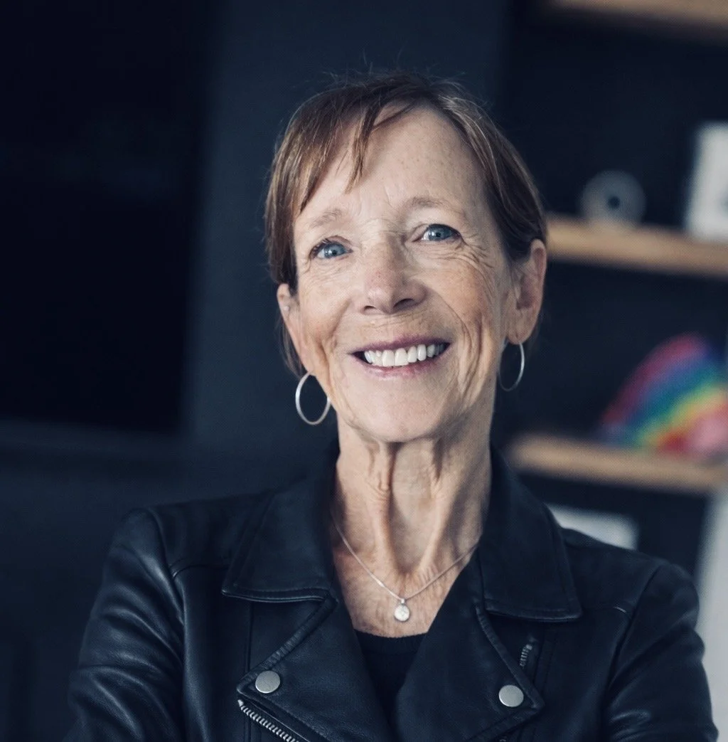 A smiling middle-aged woman with short brown hair, wearing hoop earrings, a silver necklace with a pendant, and a black leather jacket, in an indoor setting.