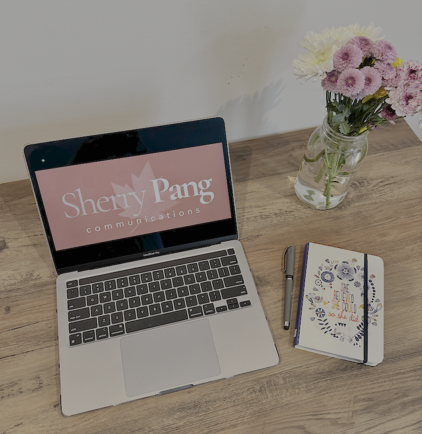 Laptop, notebook with colorful design, pen, and glass jar with pink and purple flowers on a wooden table.