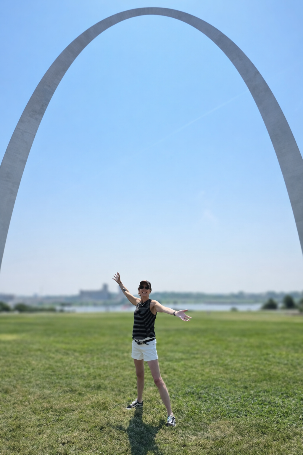 A woman standing on green grass with arms outstretched in front of the Gateway Arch in St. Louis, Missouri, on a sunny day with a clear blue sky.