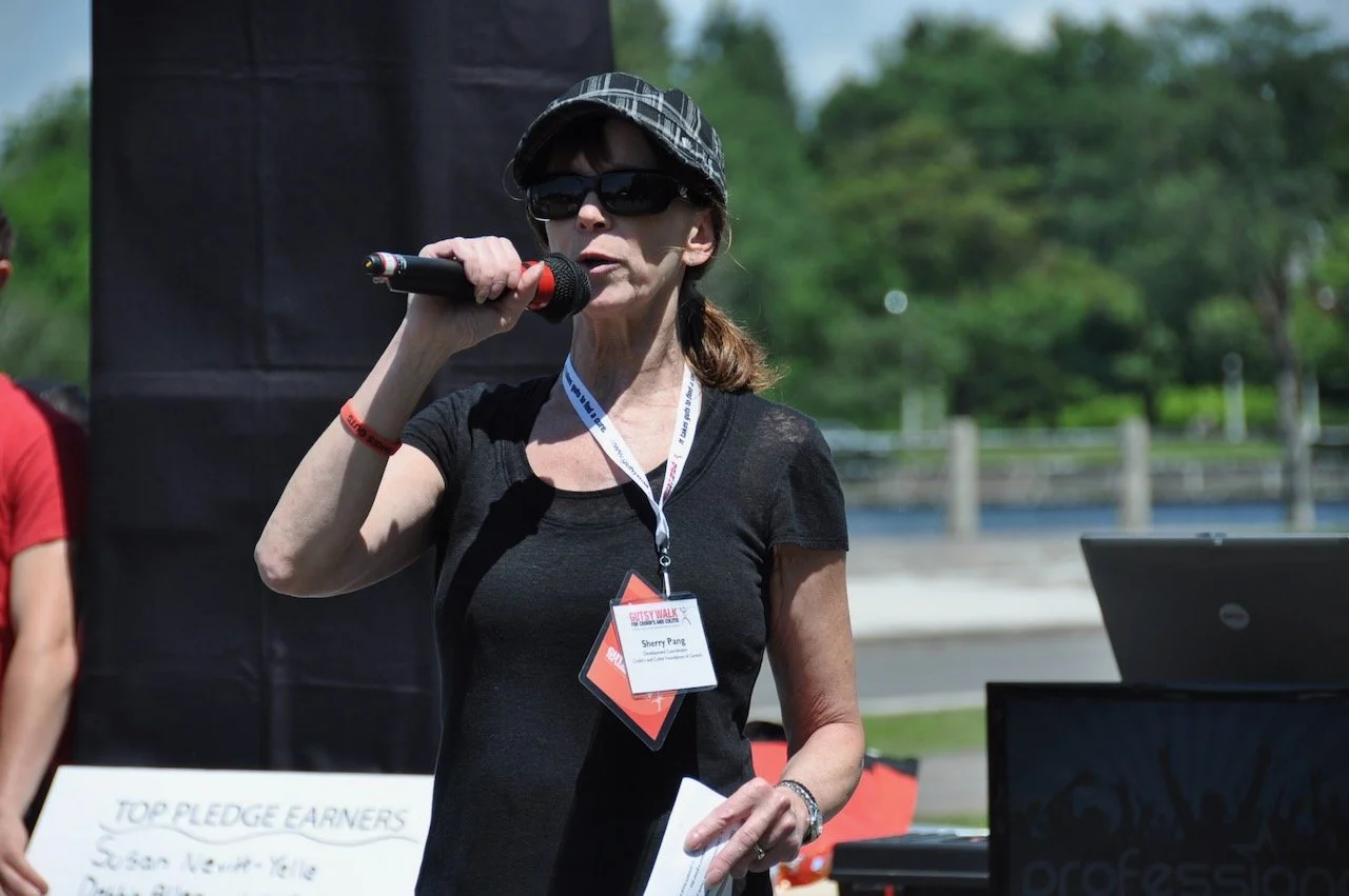 A woman wearing sunglasses, a plaid cap, and a black t-shirt speaking into a microphone at an outdoor event with trees and a body of water in the background.