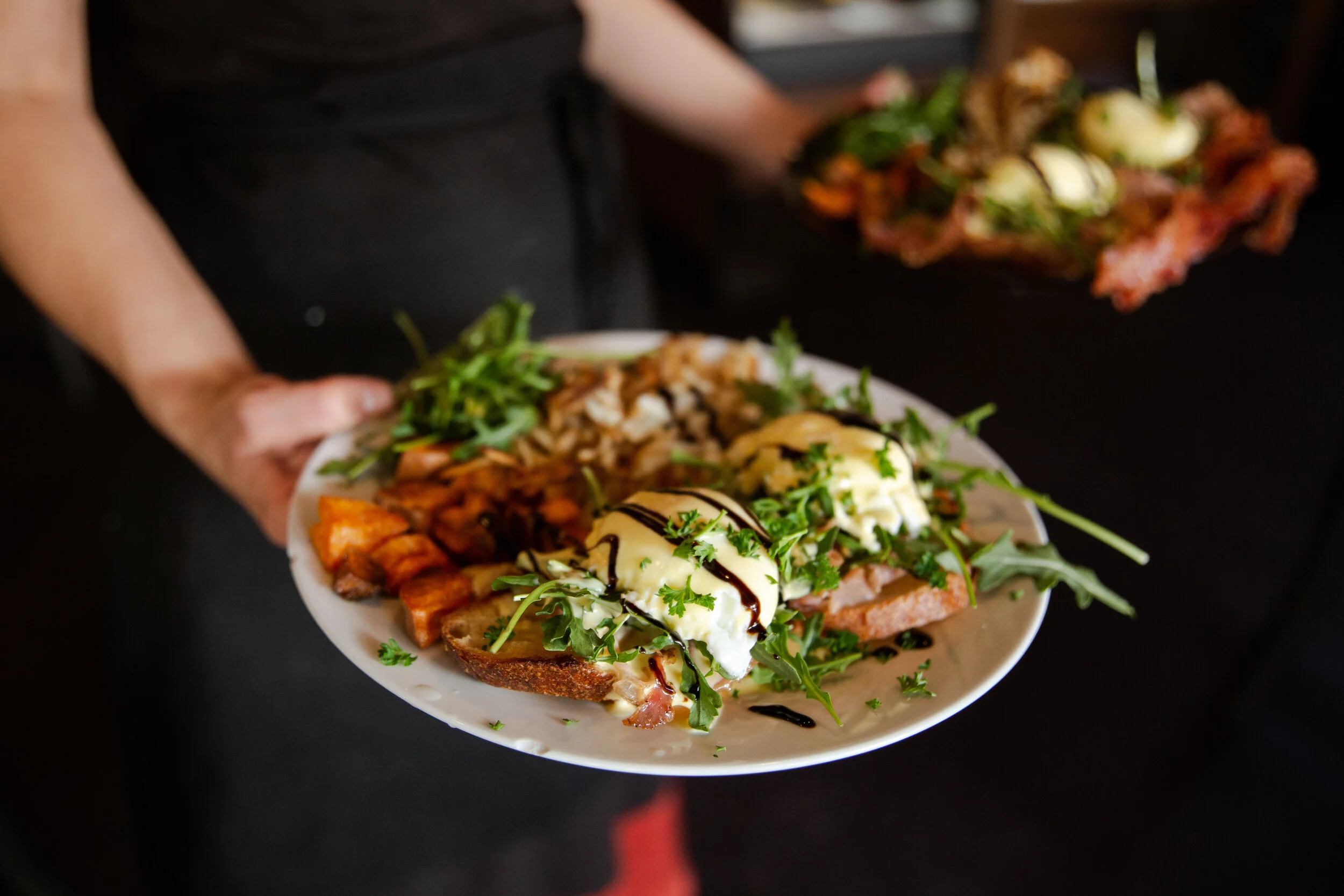 A person holding a white plate of various loaded baked potato toppings, including sour cream, shredded cheese, bacon bits, chives, and greens, with another similar plate visible in the background.