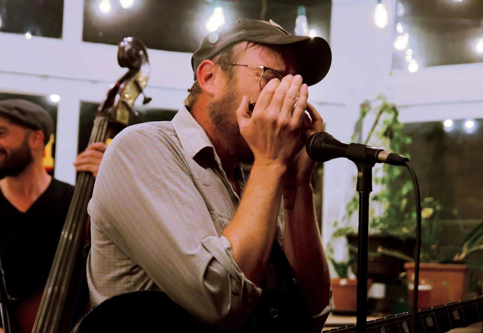 A man wearing glasses and a cap, playing harmonica near a microphone, with a man in the background holding a double bass, in a cozy indoor setting with string lights and plants.