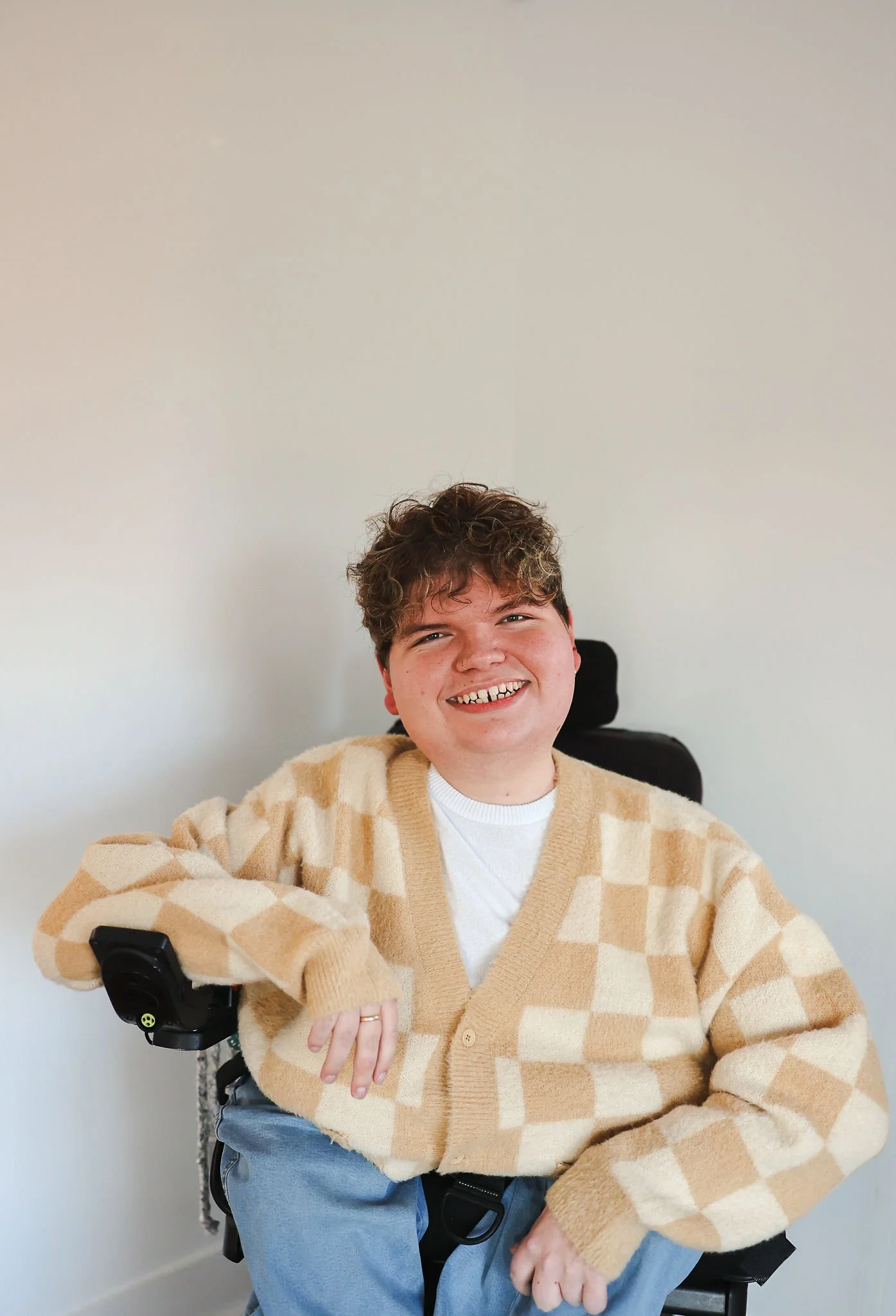 A smiling male with curly hair sitting in a motorized wheelchair, wearing a beige checkered cardigan over a white shirt, in a room with plain white walls.