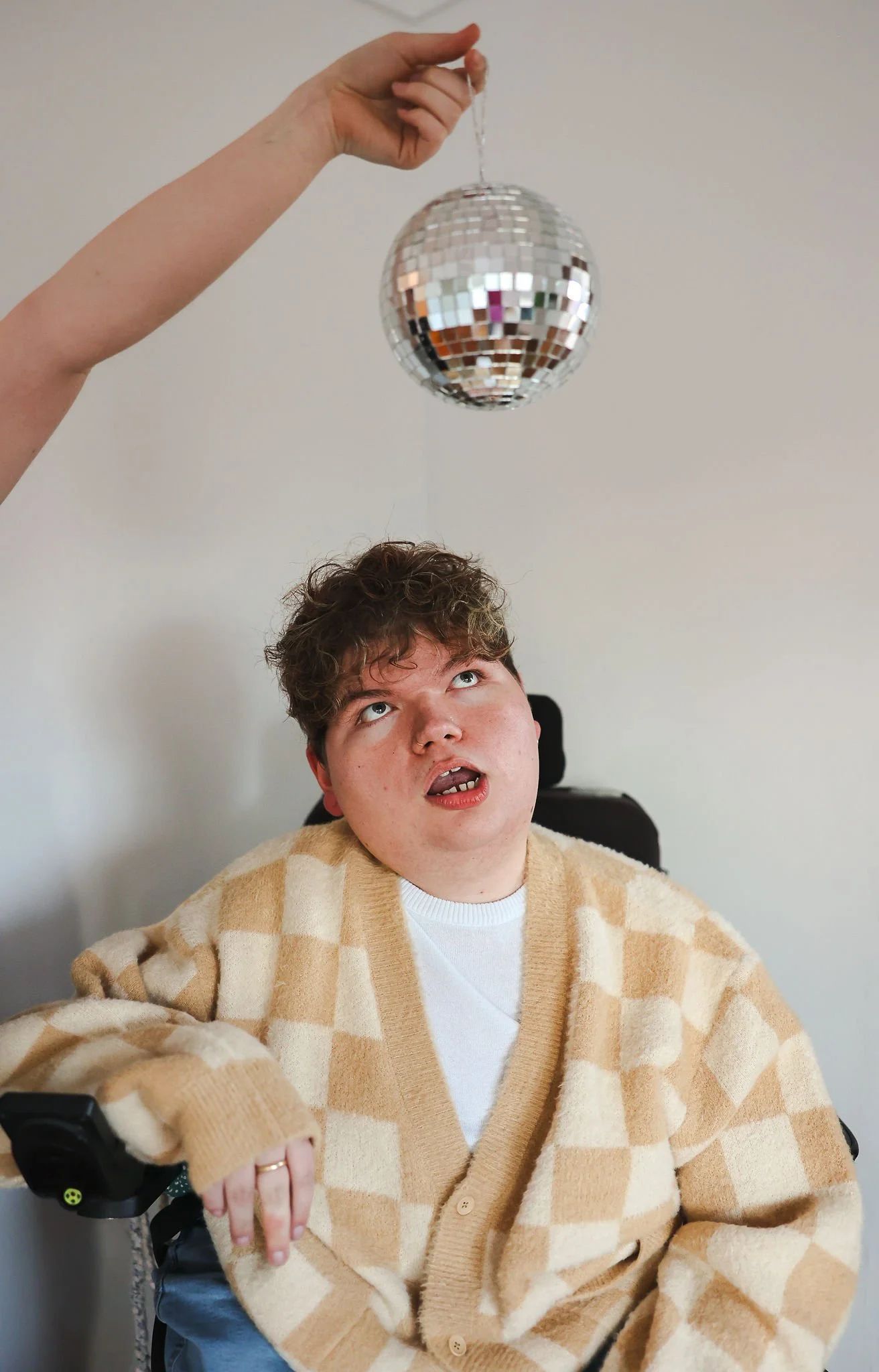 A male with curly hair and a checkered beige cardigan sitting in a wheelchair, looking up with an exaggerated expression at a disco ball being held above his head by an unseen person.
