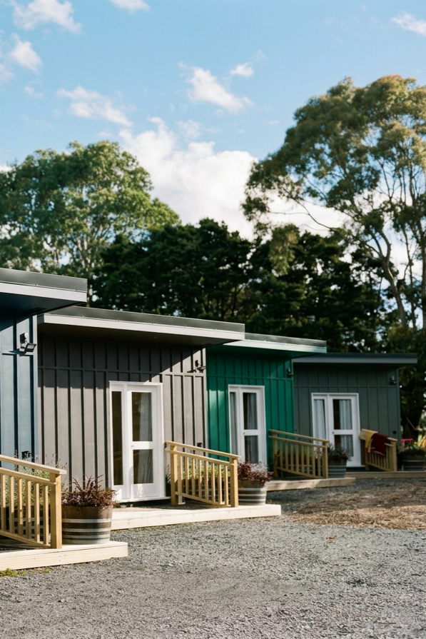 A row of small, modern cottage-style homes with colorful exteriors in shades of gray and teal, each with a small porch and potted plants, set against a backdrop of tall trees and a blue sky with some clouds.