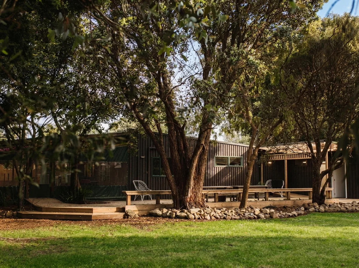 A backyard scene featuring a large tree with multiple trunks, a raised wooden deck with built-in benches and chairs, and a modern black wooden venue in the background. There is a well-maintained green lawn in the foreground.