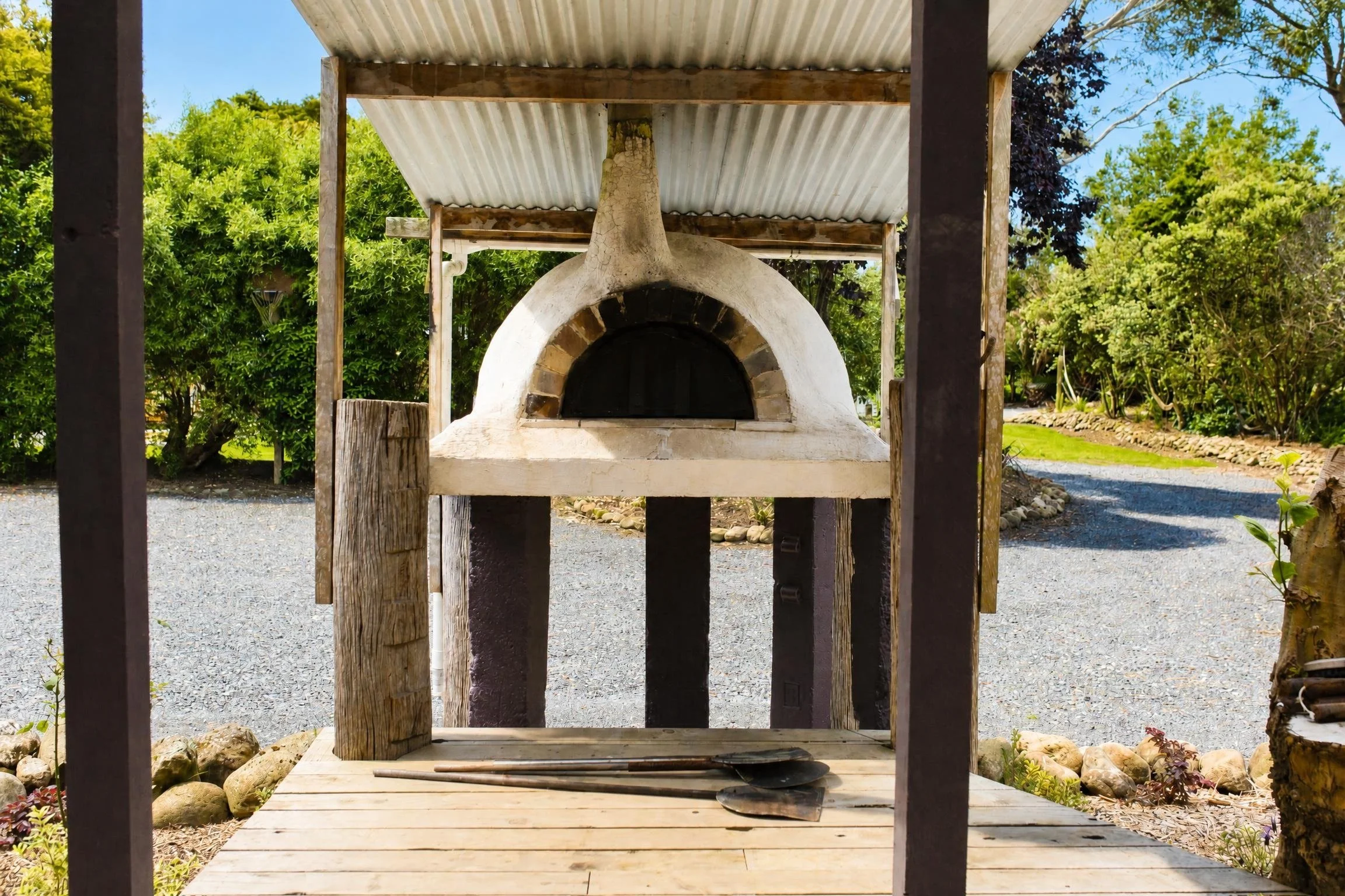 Outdoor pizza oven with a wooden platform, a metal roof, surrounded by greenery.