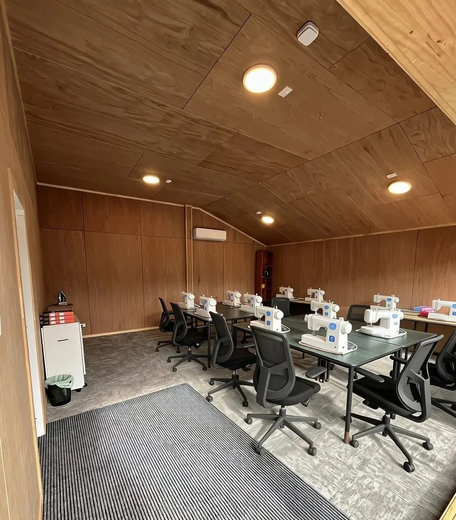 A room with wooden walls and ceiling, equipped with multiple sewing machines on tables, black office chairs, and a small cabinet with red and black boxes, suggesting a sewing or crafting class.