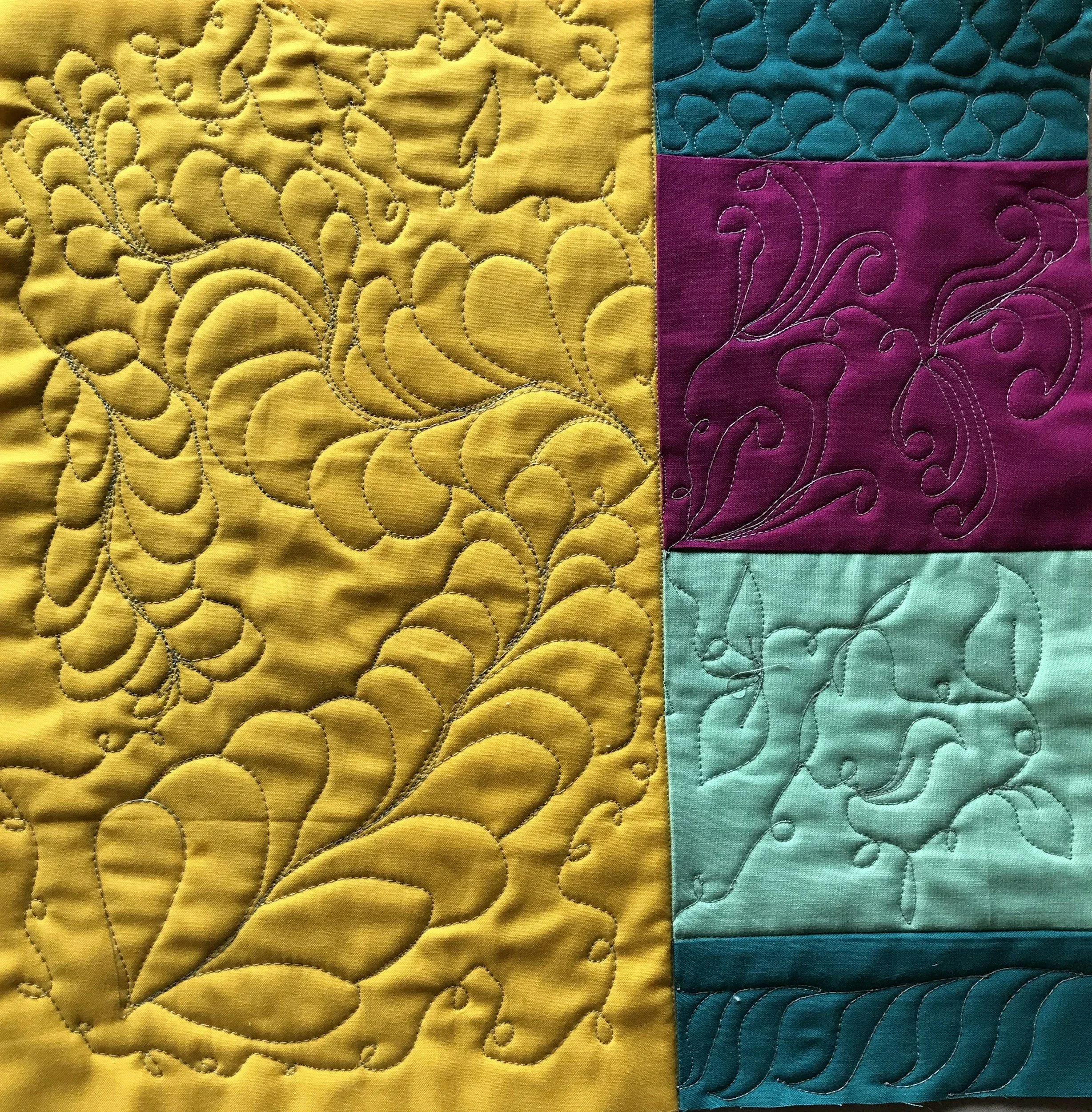 Close-up of a patchwork quilt with yellow, teal, and purple sections, featuring intricate multicolored stitching and quilted patterns.