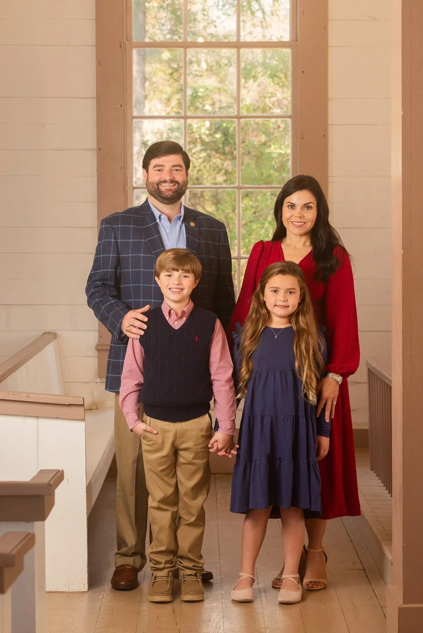 A family of four standing inside a house near a large window with trees outside. The father wears a checkered blazer and light blue shirt, the mother wears a red dress, the boy wears a pink shirt with a navy vest, and the girl wears a navy dress with light-colored shoes.