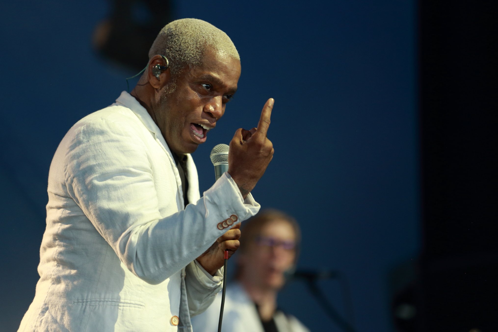 A person with short, bleach blonde hair, wearing a white jacket, speaking into a microphone, raising one finger, with another person blurred in the background.
