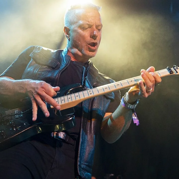 A man playing an electric guitar on stage, wearing a black leather vest, with stage lighting creating a smoky background.