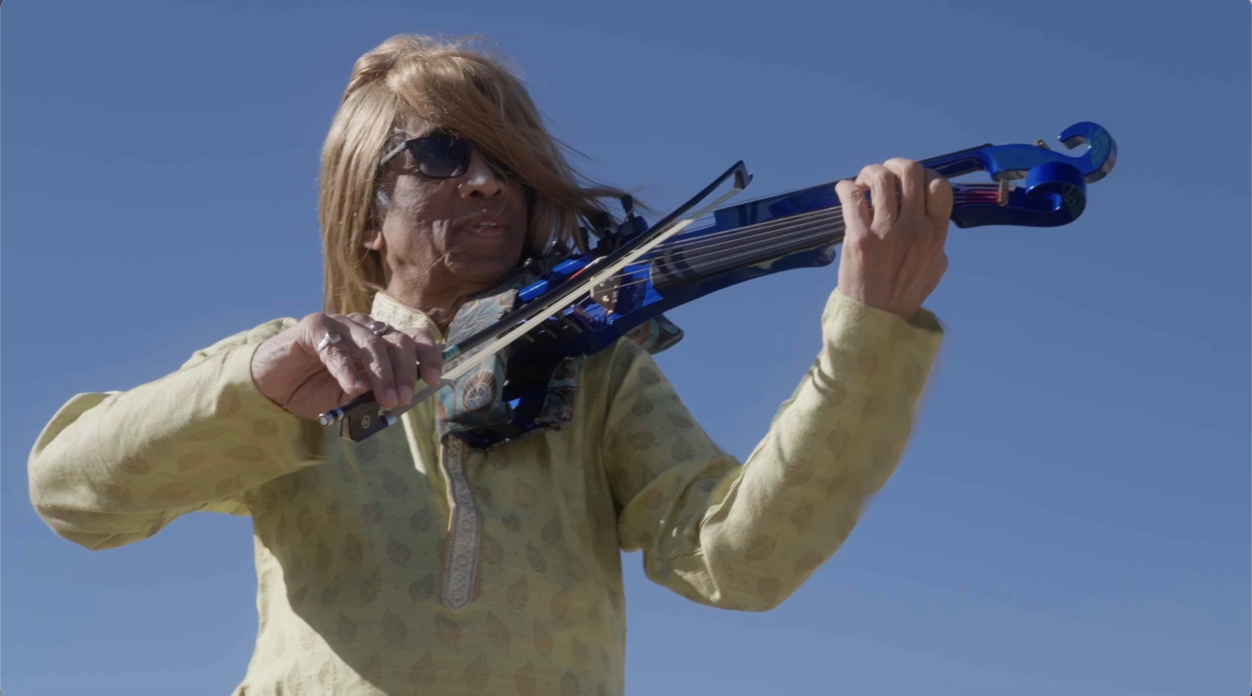 Older man with long, blond hair, wearing sunglasses and a yellow patterned shirt, playing a blue electric violin outdoors against a clear blue sky.