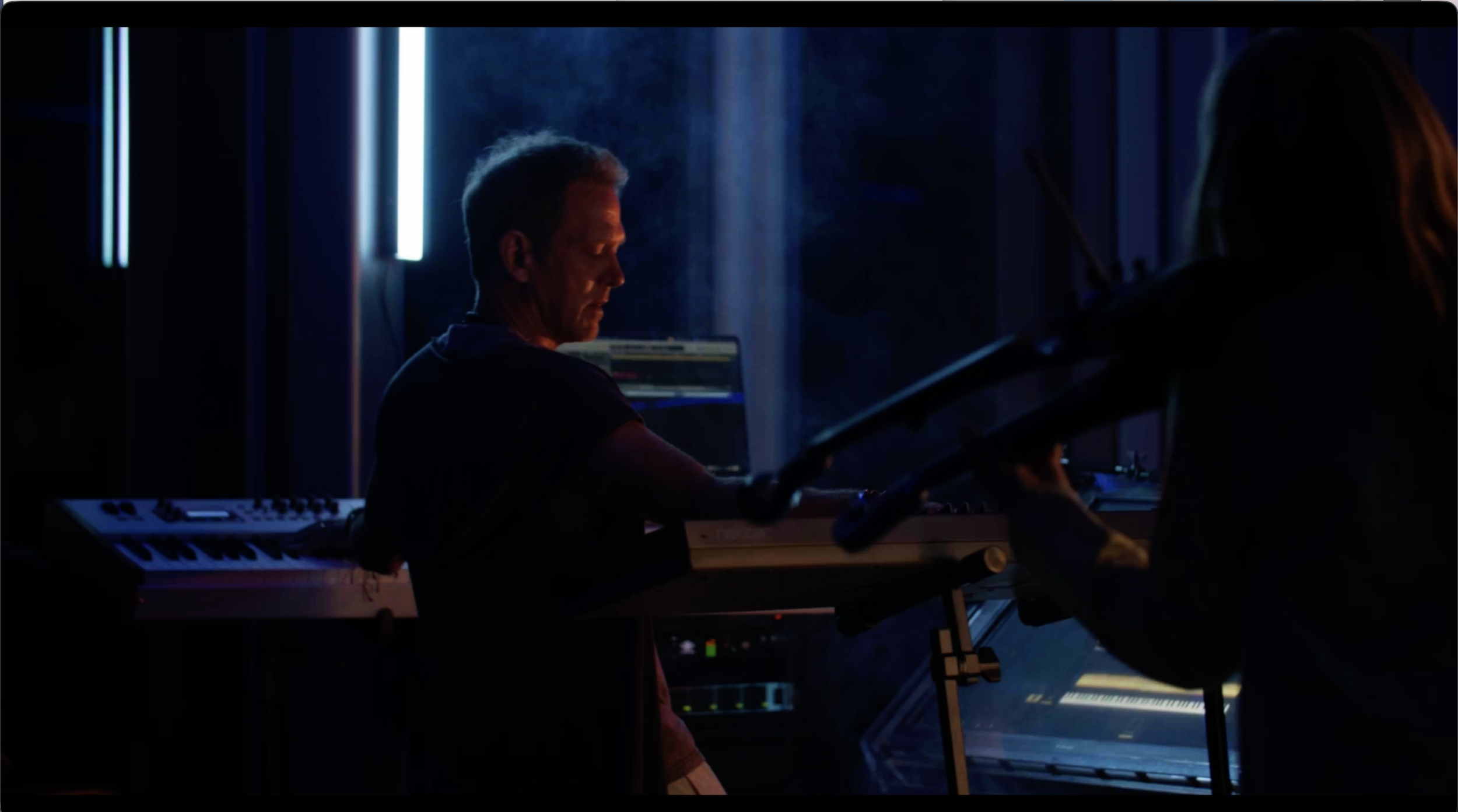 Two musicians are performing in a dimly lit studio, playing keyboards and a violin, with blue and purple lighting creating a moody atmosphere.