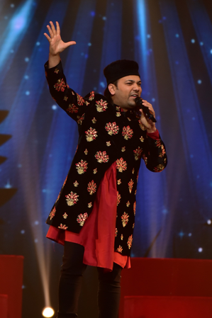 Male comedian wearing a black kurta with floral patterns, red kurta underneath, and black cap, performing on stage with a microphone, raising his right hand, with a starry blue background.