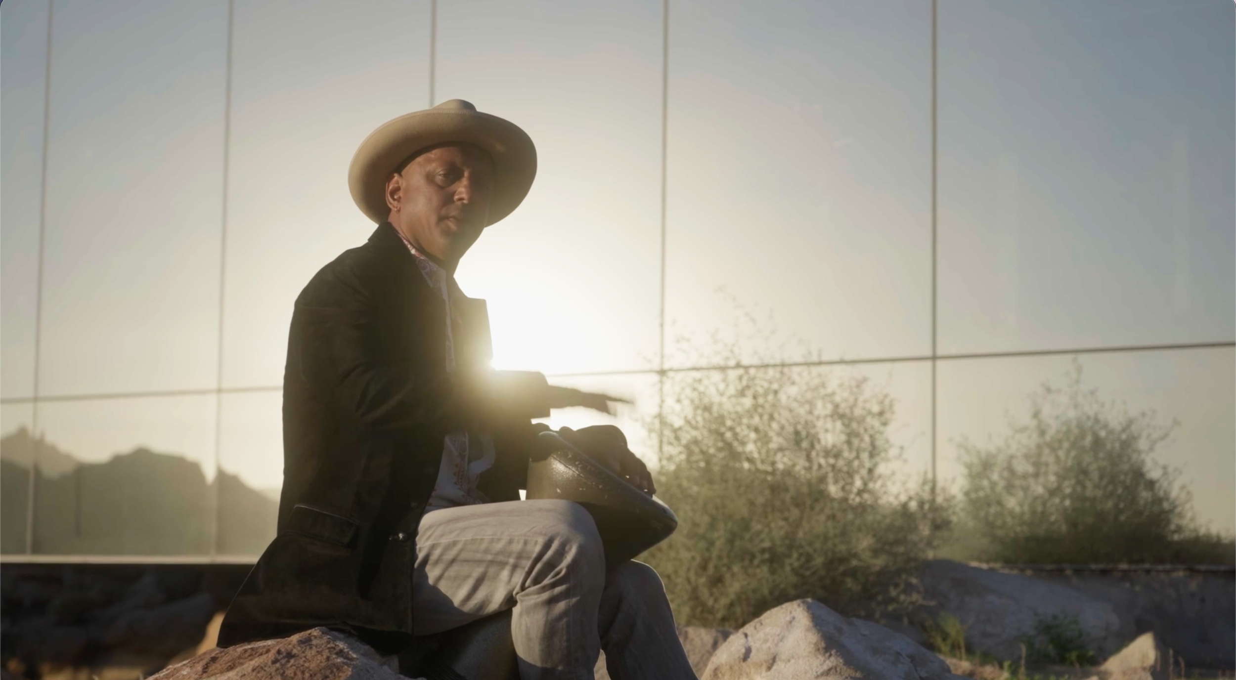 A person sitting on rocks, wearing a wide-brimmed hat and a dark jacket, holding a metallic bowl, with sunlight behind them and a reflective building wall in the background.
