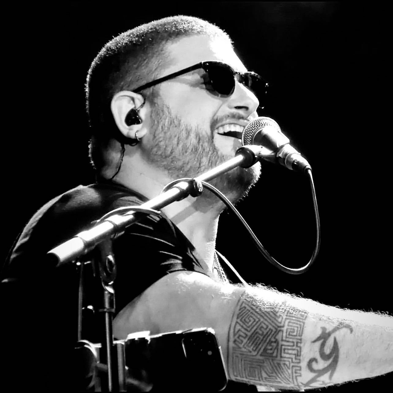 Black and white photograph of a male musician singing into a microphone, wearing sunglasses, with a microphone stand, tattoos on his arm, and an earpiece in his ear.
