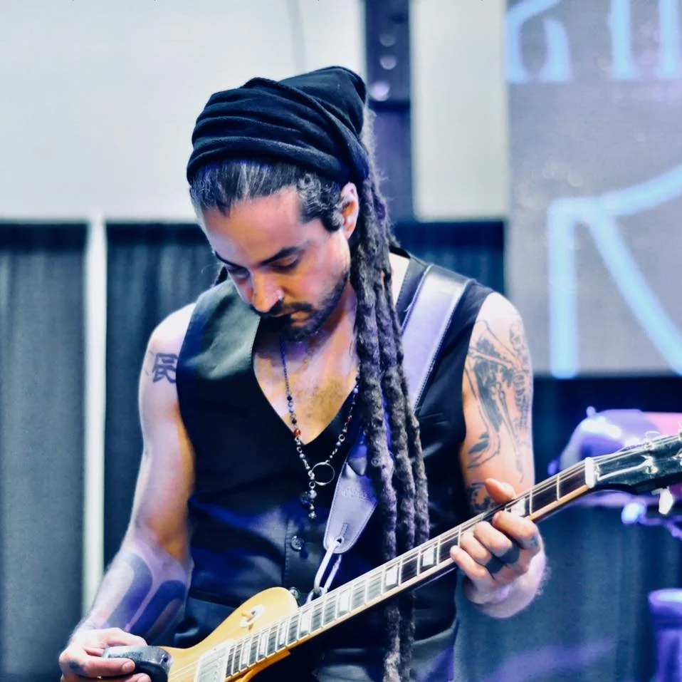 A man with long dreadlocks wearing a black sleeveless shirt and a black headwrap, playing an electric guitar in a performance setting.
