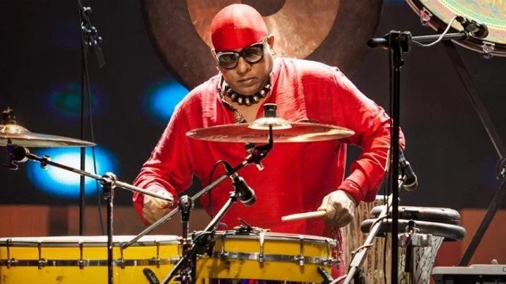 A musician dressed in a red shirt and red head covering playing drums on stage.