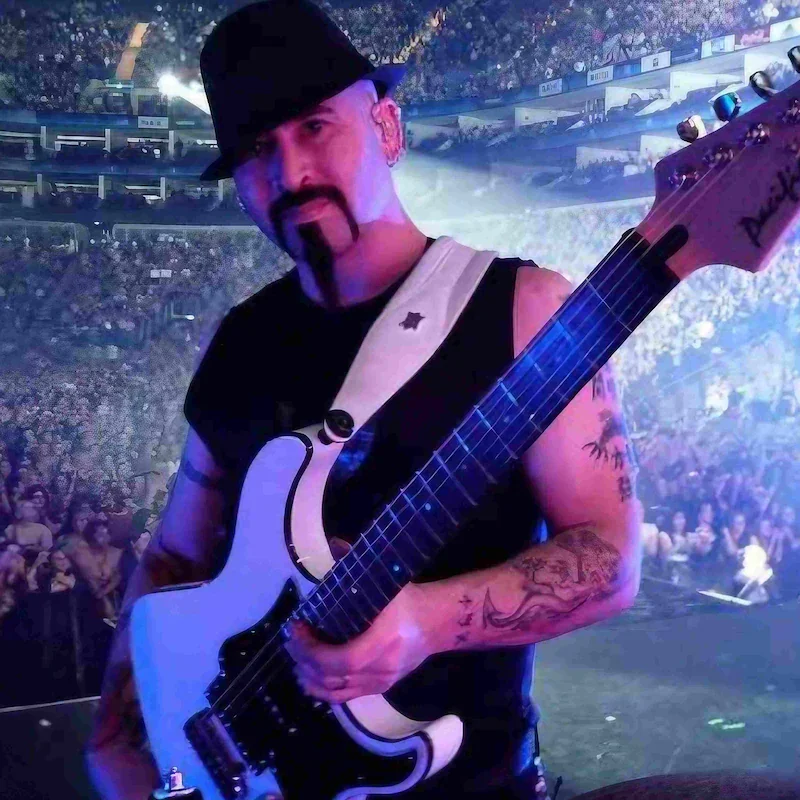 Musician on stage holding an electric guitar with tattoos on his arms at a concert in a large arena.